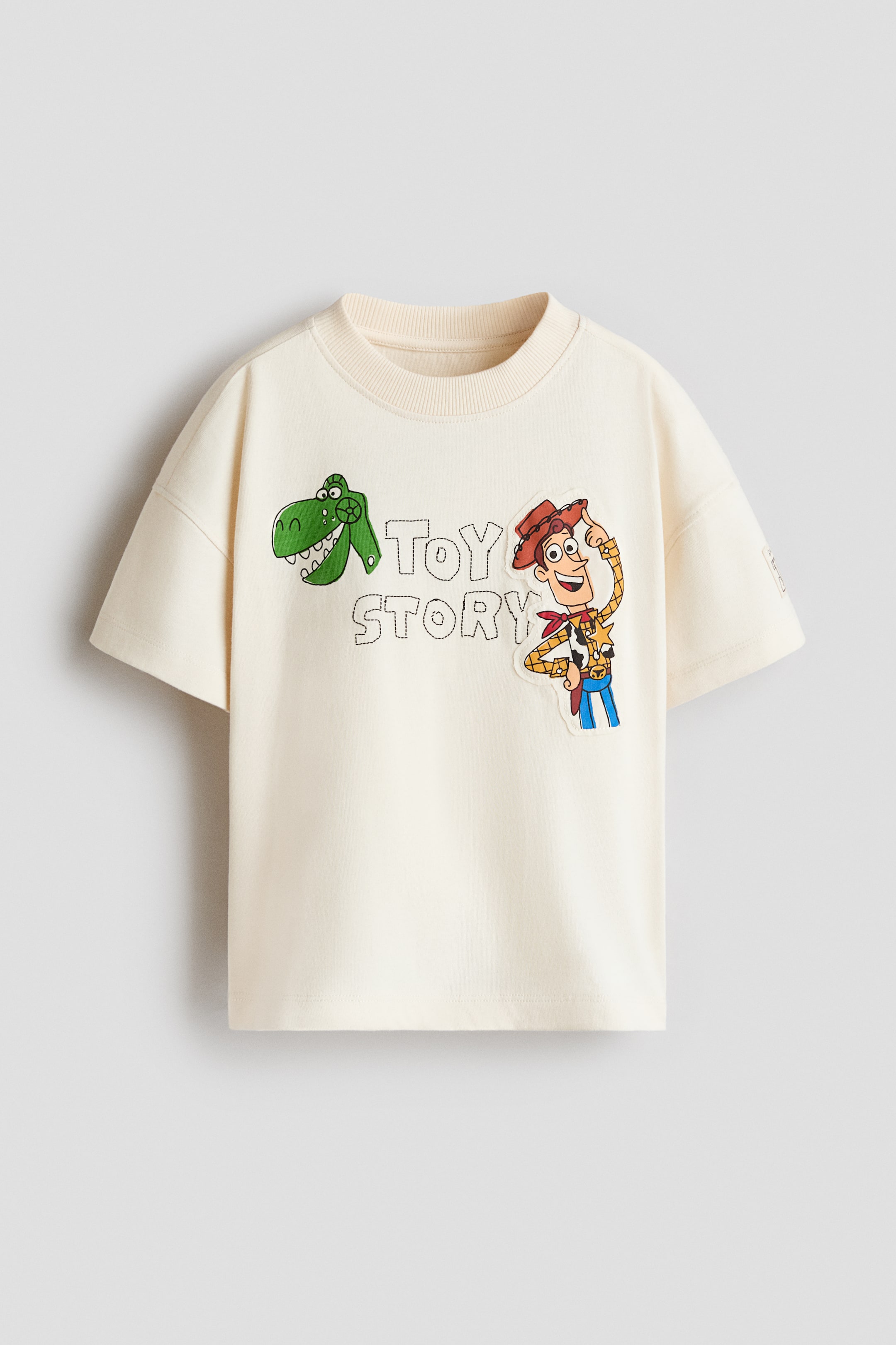 Größeres Bild anzeigen: Ein oversized T-Shirt in Creme wird von vorne gezeigt und verfügt über überschnittene Schultern sowie einen Rippstrick-Ausschnitt. Auf der Brust ist ein Grafikdesign mit den Figuren Rex und Woody zu sehen, die den im Stickstil gehaltenen Schriftzug „TOY STORY“ umrahmen.