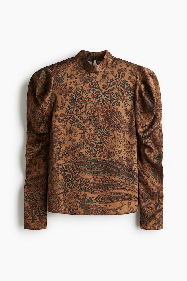 Eine Vorderansicht einer Bluse zeigt eine goldbraune Basis mit einem komplexen Paisleymuster in Schattierungen von Dunkelbraun, Tealgrün und Burgunderrot. Sie ist mit einem Stehkragen und voluminösen, gepufften langen Ärmeln gestaltet, die zu den Handgelenken hin schmaler werden. - 1