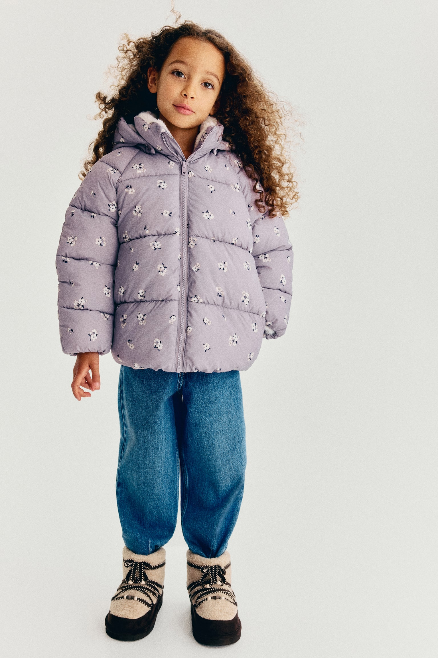 A childâs light purple puffer jacket with a small, scattered white floral print is shown from the front, featuring horizontal quilting and a full-length zip closure. The jacket also shows an attached hood lined with soft, cream-coloured fleece. - 1