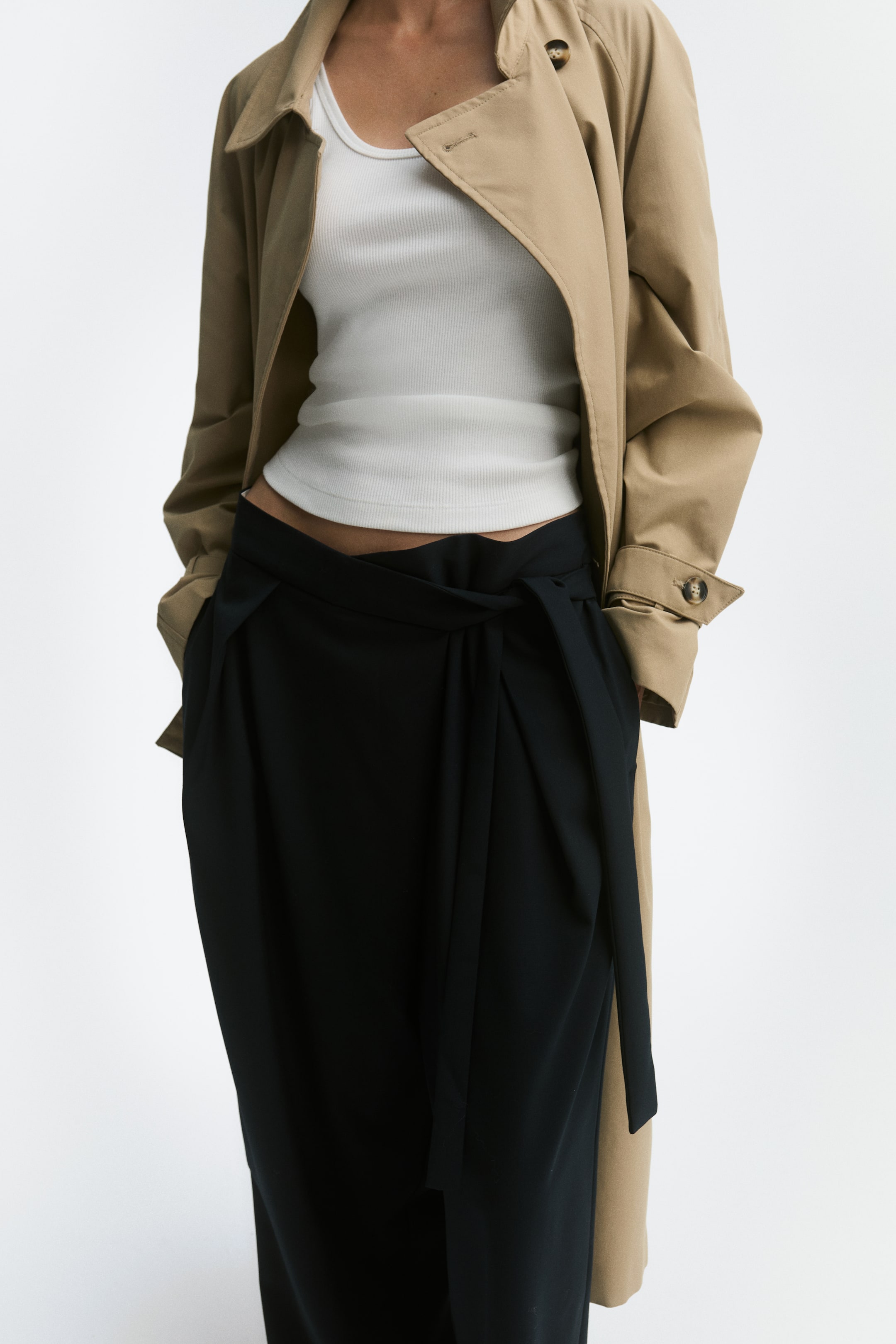 Voir une image plus grande: Ce pantalon noir coupe ample présente une taille haute avec un devant croisé et une fermeture à nouer sur un côté, ainsi que des plis à l’avant qui tombent en une silhouette à jambe ballon. Le pantalon est présenté de face, agencé avec un haut sans manches côtelé blanc et un trench beige ouvert.