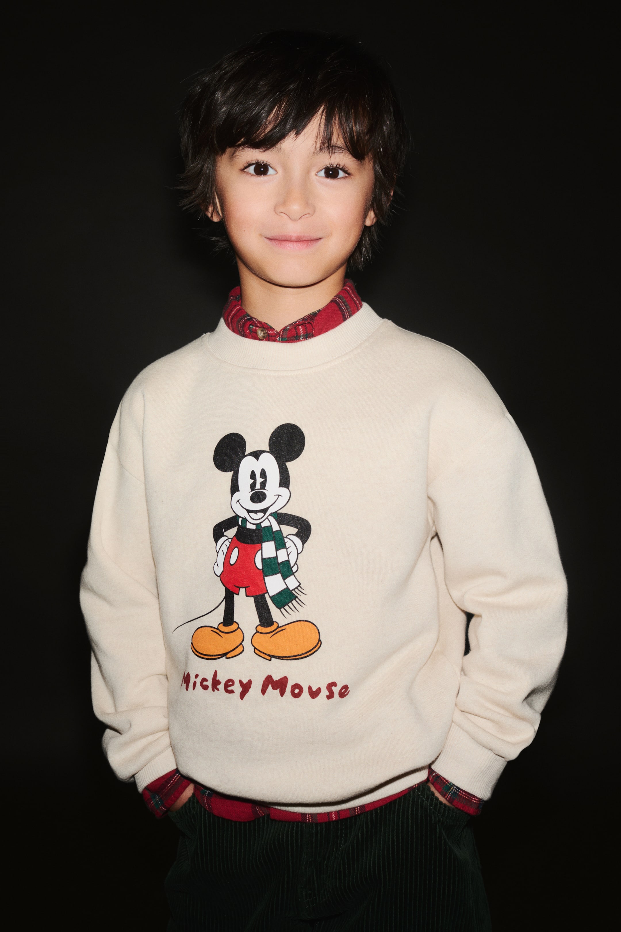 Agrandir l'image: Un sweat-shirt en polaire de couleur crème est présenté de face, avec un motif imprimé de Mickey Mouse et le texte « Mickey Mouse » inscrit en dessous. Le sweat est doté d’un col rond en bord-côte, d’épaules tombantes et de poignets côtelés, et est porté superposé sur une chemise à col à carreaux rouges.