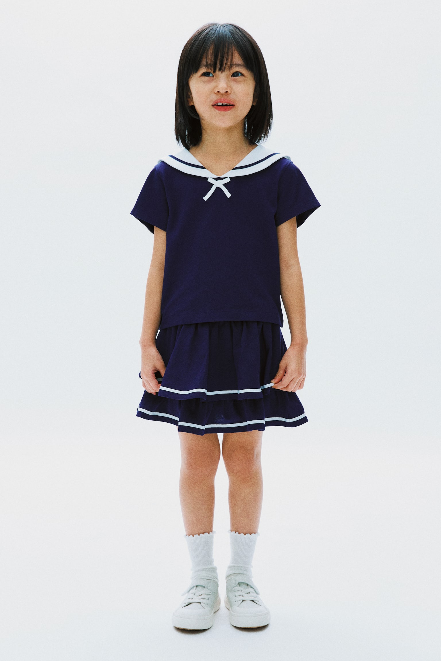 A navy blue two-piece set, composed of a short-sleeved top featuring a white sailor collar and a white bow accent, paired with a tiered skirt adorned with light blue horizontal stripes, is showcased by the model. - 1