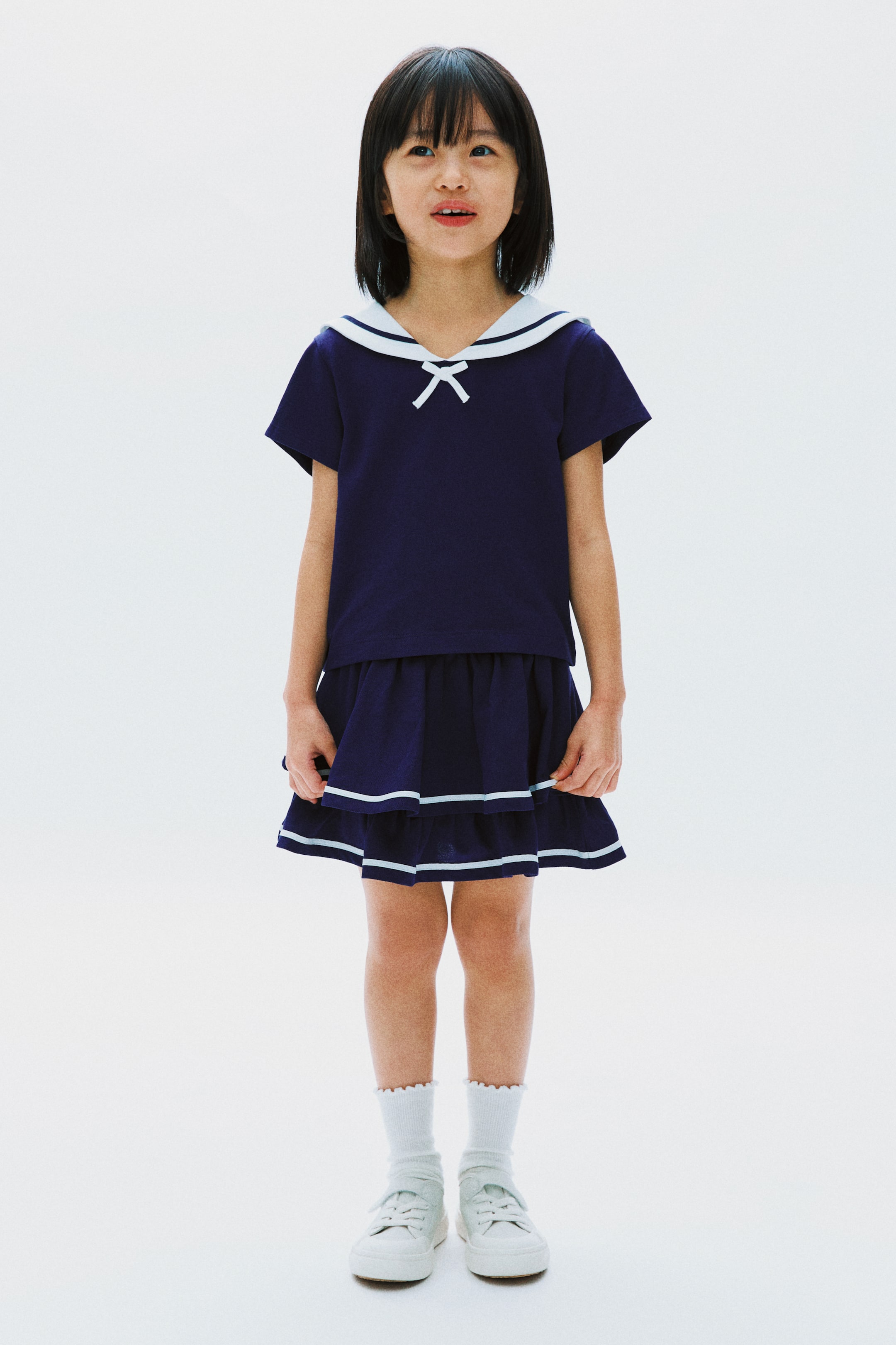 View larger image: Model tersebut memakai set dua helai berwarna biru navy, terdiri daripada baju pendek berlengan dengan kolar pelaut putih dan perincian reben putih, serta skirt berlapis dengan jalur mendatar biru muda.