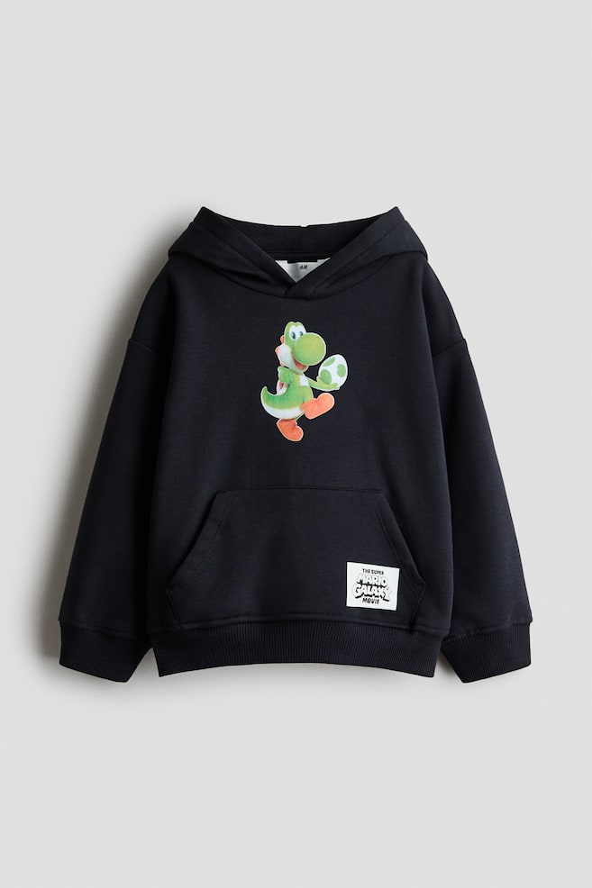 Una felpa con cappuccio nera dal fit morbido Ã¨ mostrata frontalmente, con una grafica stampata di Yoshi che tiene un uovo maculato al centro del petto. Ã caratterizzata da un cappuccio con davanti incrociato, spalle scese, una tasca a marsupio frontale con un piccolo patch bianco e polsini e orlo a coste. - 1