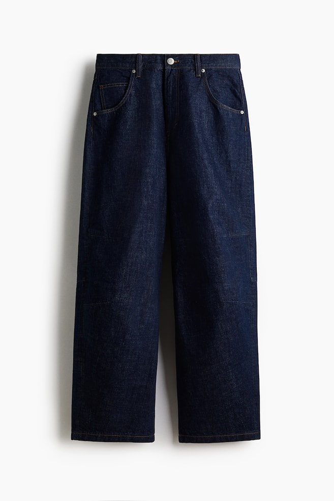 Eine dunkelblaue, weit geschnittene Jeans wird von vorne präsentiert, mit einem Knopf- und Reißverschluss, fünf Gürtelschlaufen und zwei geschwungenen Fronttaschen mit Nieten-Details, einschließlich einer kleinen Münztasche. Kontrastierende Steppnähte sind überall sichtbar, und zwei rechteckige Knie-Patches sind auf der Vorderseite jedes Beins aufgenäht. - 1