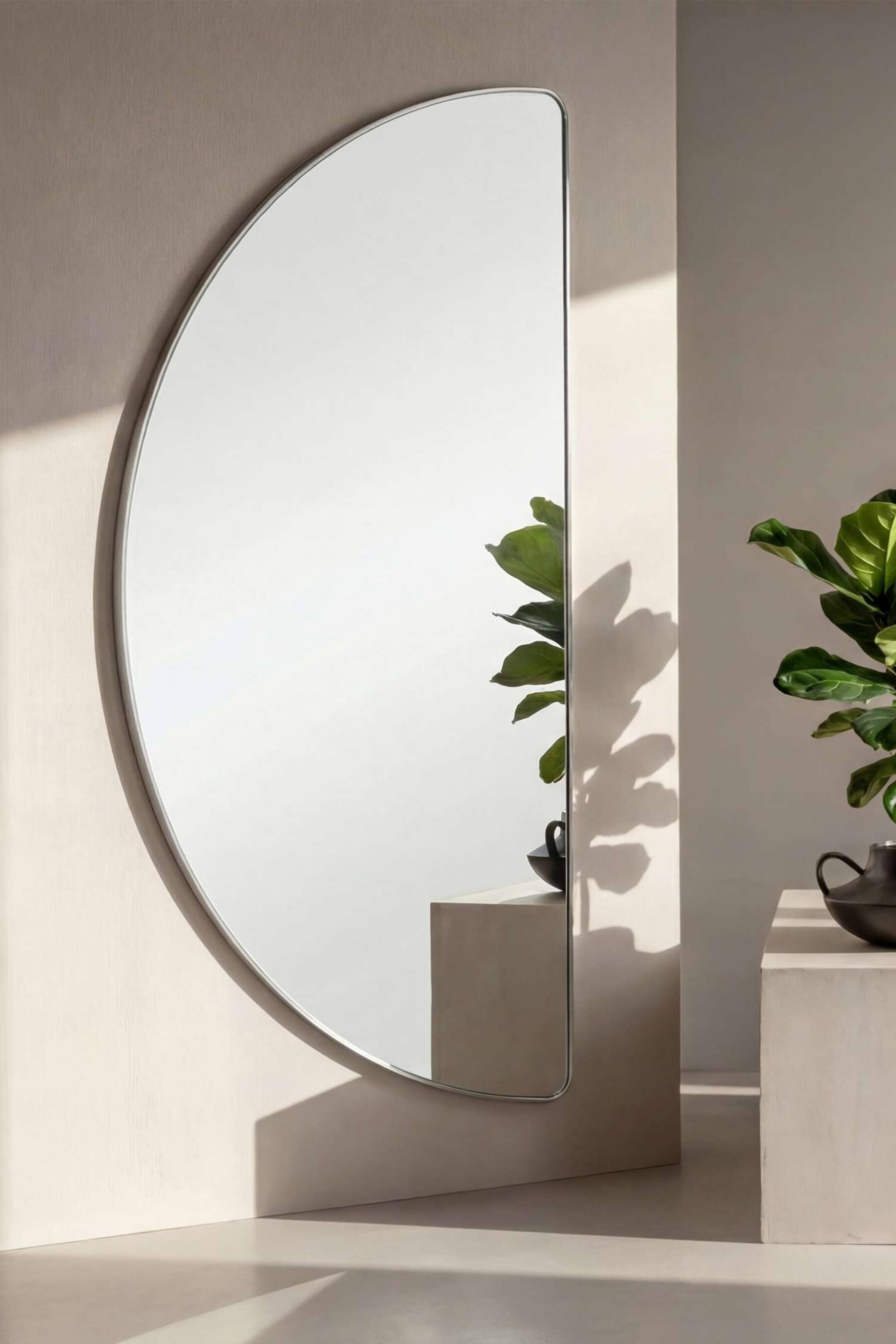 Se større billede: Mona Large Mirror hænger på en væg og fremviser en halvcirkelform med en flad lodret kant på højre side og en slank, sølvtonet ramme. Spejlet reflekterer en nærliggende potteplante og en piedestal og demonstrerer dermed sin klare, reflekterende glasoverflade.