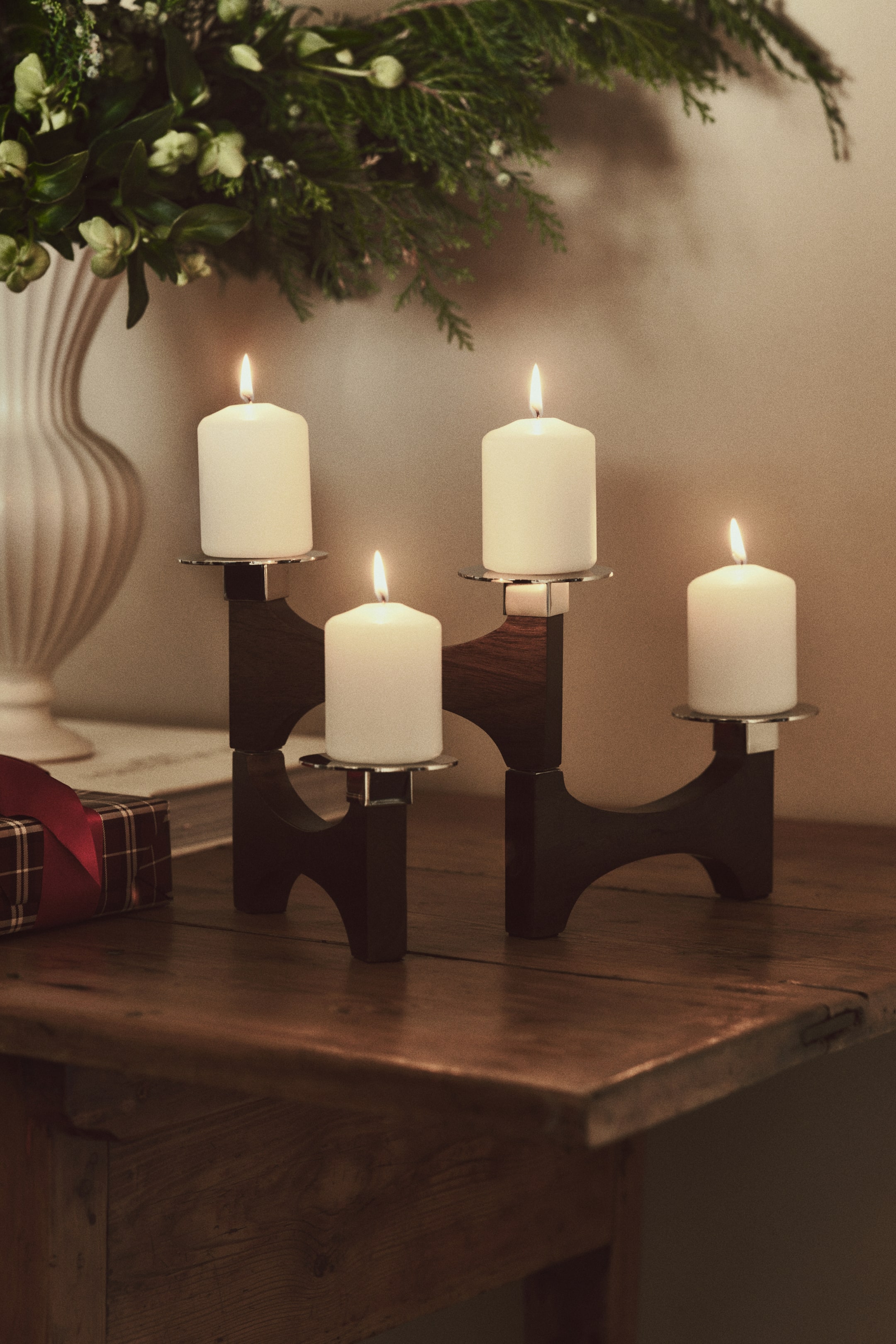 View larger image: A foldable candelabra in dark mango wood, facing front, displays a multi-level structure of interlocking curved wooden pieces. The candelabra holds four lit white pillar candles on its four stainless steel plate holders.