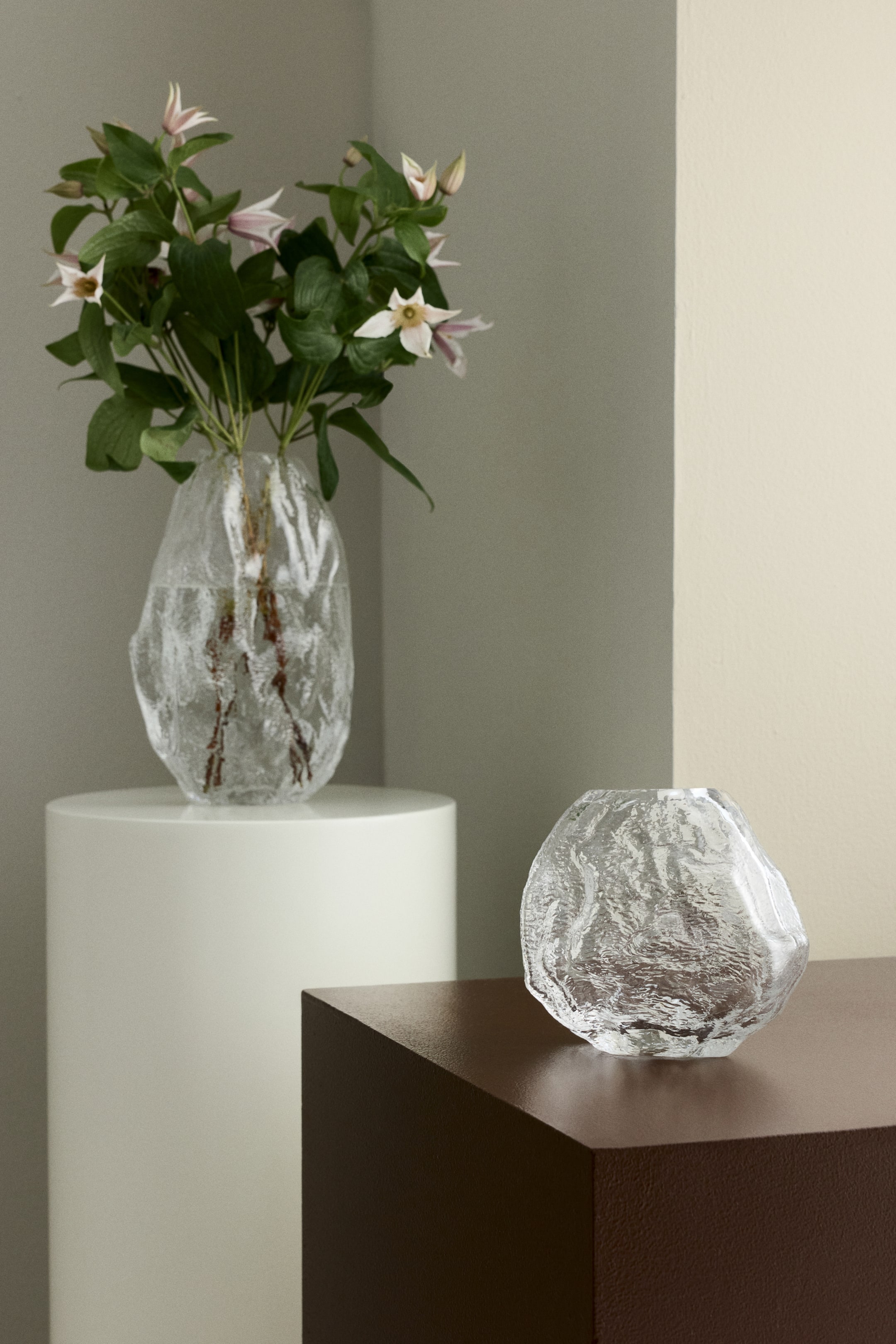 Agrandir l'image: Deux vases en verre transparent présentent des formes irrégulières et organiques, avec une surface texturée évoquant la glace. Le vase le plus grand contient un feuillage vert agrémenté de fleurs rose pâle et blanches, tandis que le vase le plus petit est vide.
