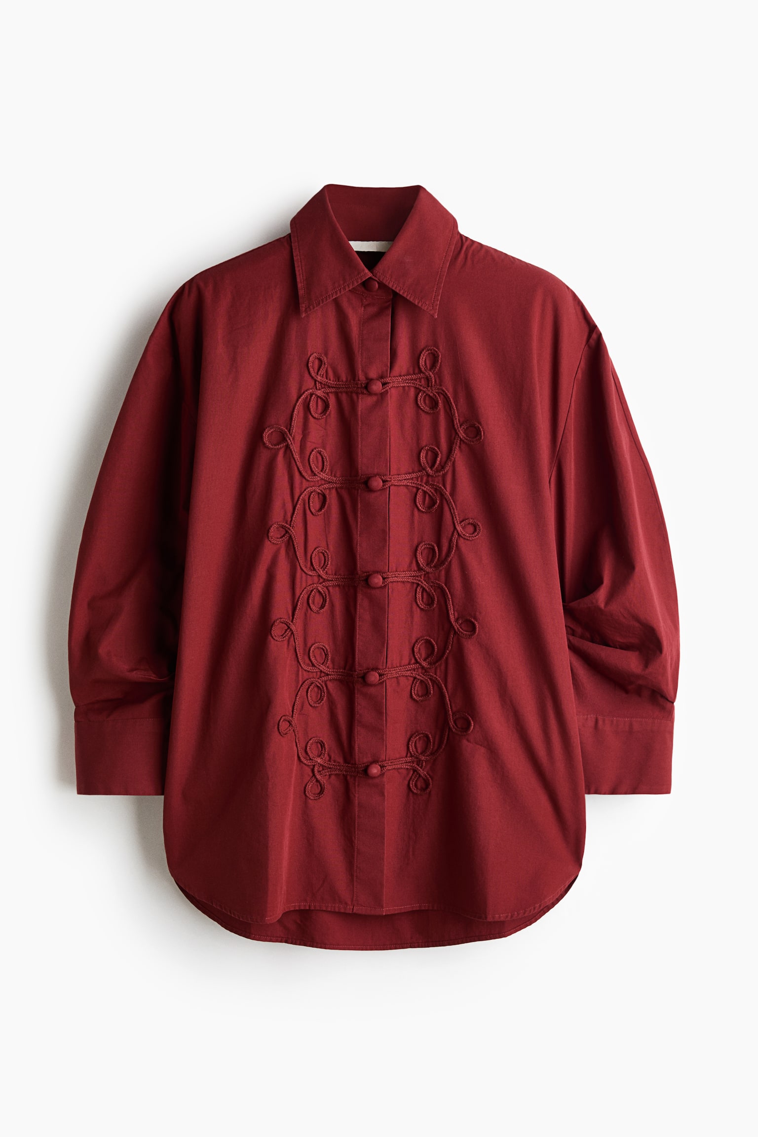 An oversized dark red cotton poplin shirt is shown from the front, featuring a point collar, dropped shoulders, and wide-cuffed sleeves. The front placket is detailed with intricate decorative cord trims that transition into loops with covered buttons, finishing with a rounded hem.