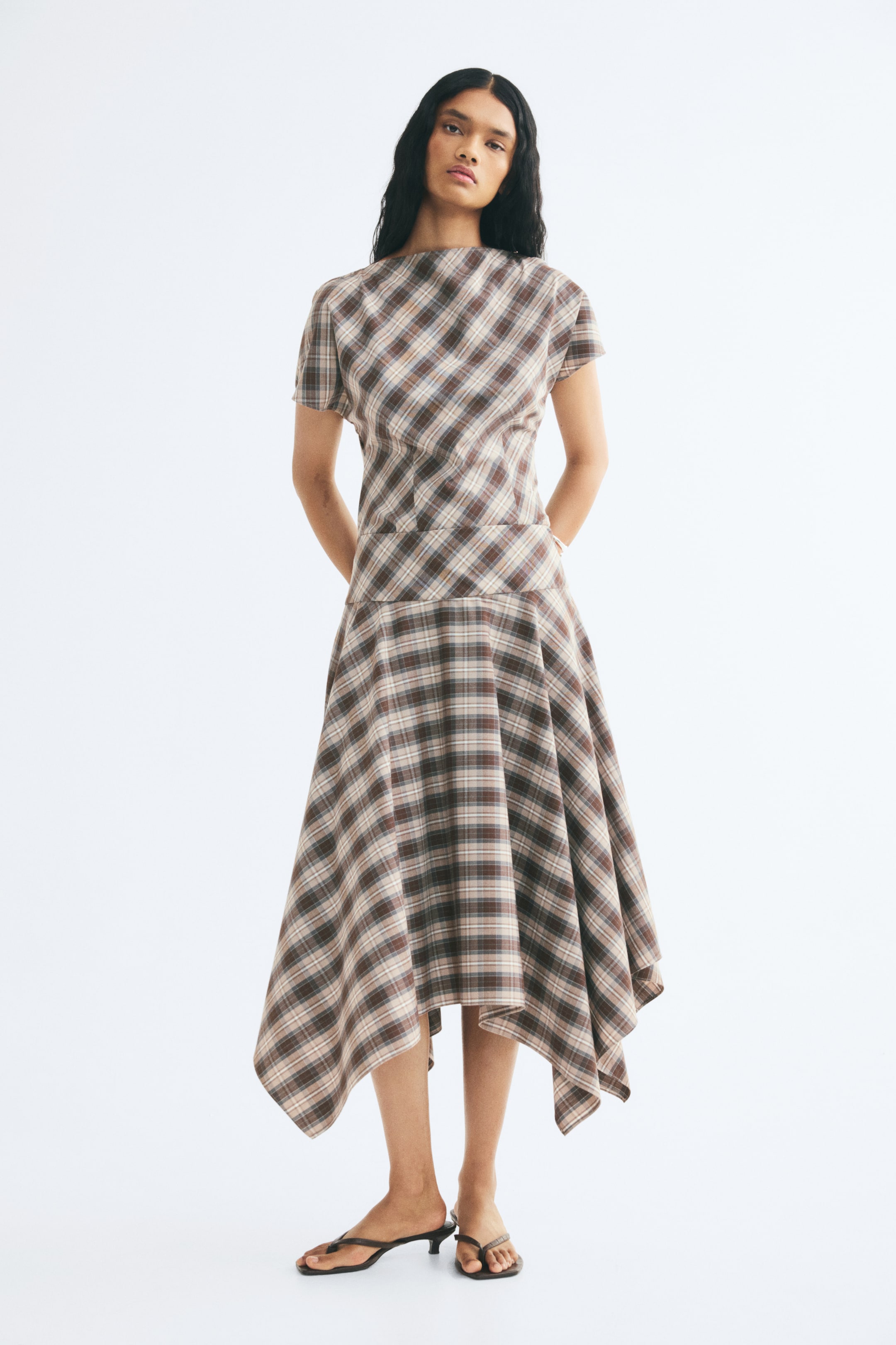 View larger image: The model is wearing a calf-length skirt in a brown and beige plaid woven fabric, featuring a wide, double-layered waistband and an asymmetrical handkerchief hem. The skirt is styled with a matching short-sleeved plaid top and low-heeled sandals.
