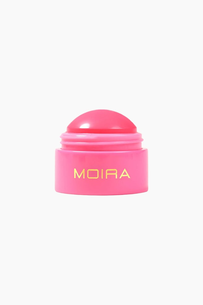 En blank, kuppelformet varm rosa kosmetisk balsam presenteres i en lys rosa sylindrisk beholder. Beholderen viser ordet "MOIRA" i gullbokstaver på forsiden, og hele produktet er vinklet litt nedover, slik at balsamens avrundede overflate vises. - 1