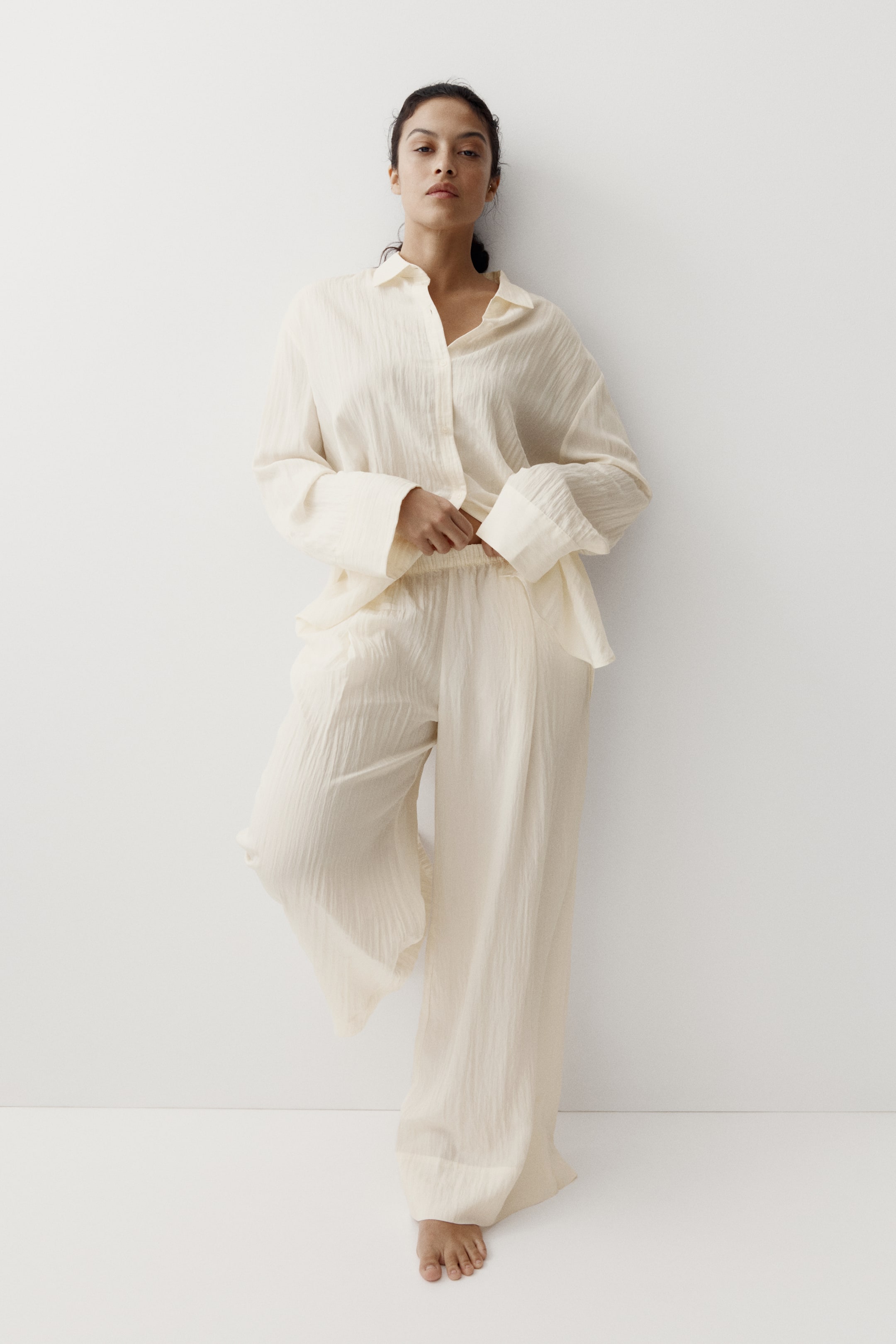 View larger image: A cream-colored, textured two-piece set is displayed from a front view. It features a collared button-down shirt with wide sleeves and buttoned cuffs, worn partially unbuttoned and tucked into wide-leg pants. The pants have an elastic waistband, and the entire set showcases a relaxed fit.