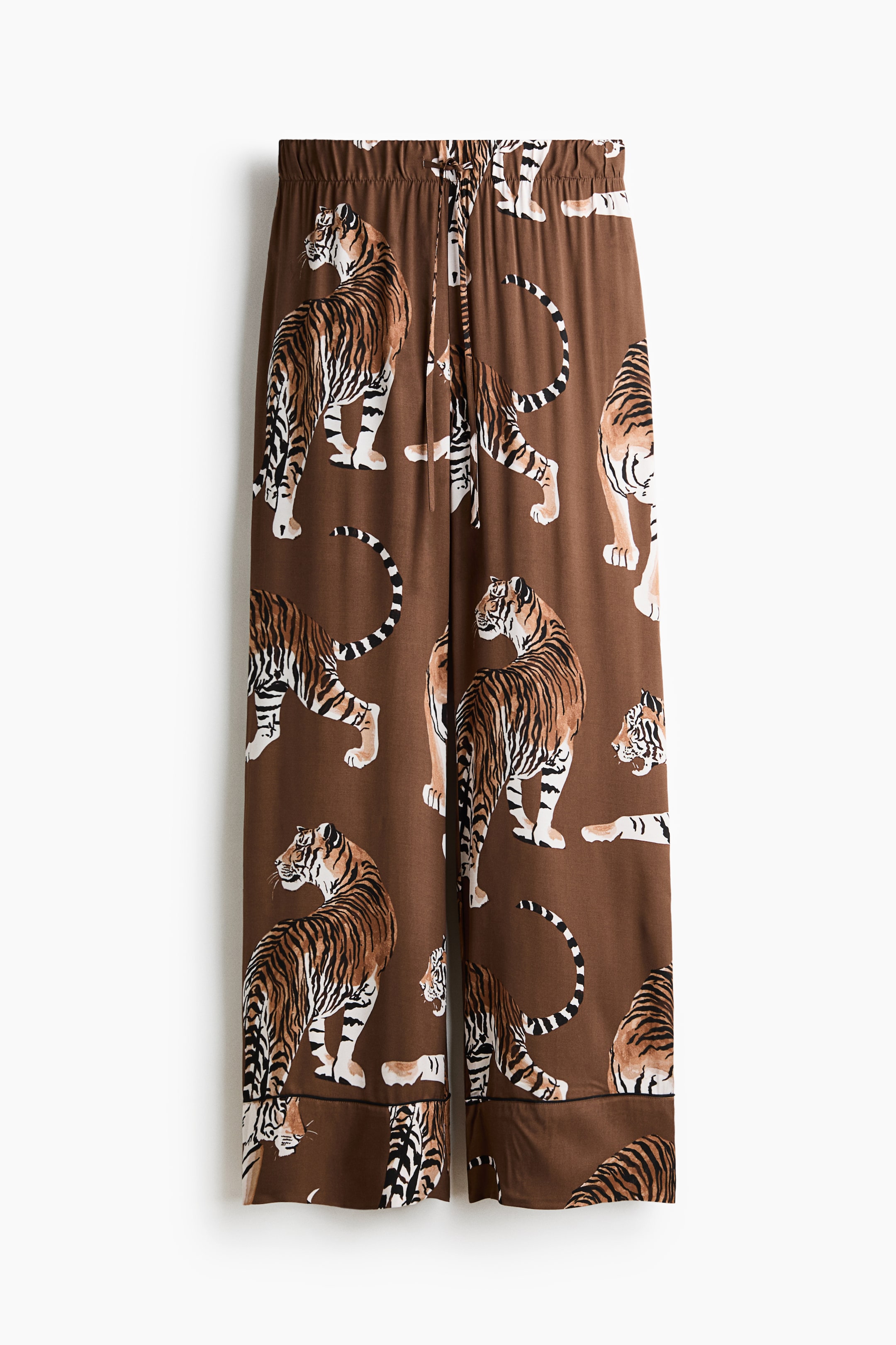 View larger image: These loose-fit pants are shown from the front, featuring a high waist with covered elastic and a narrow drawstring. They showcase a brown fabric with a repeating tiger print, draping softly into wide legs with narrow black piping just above the hems.