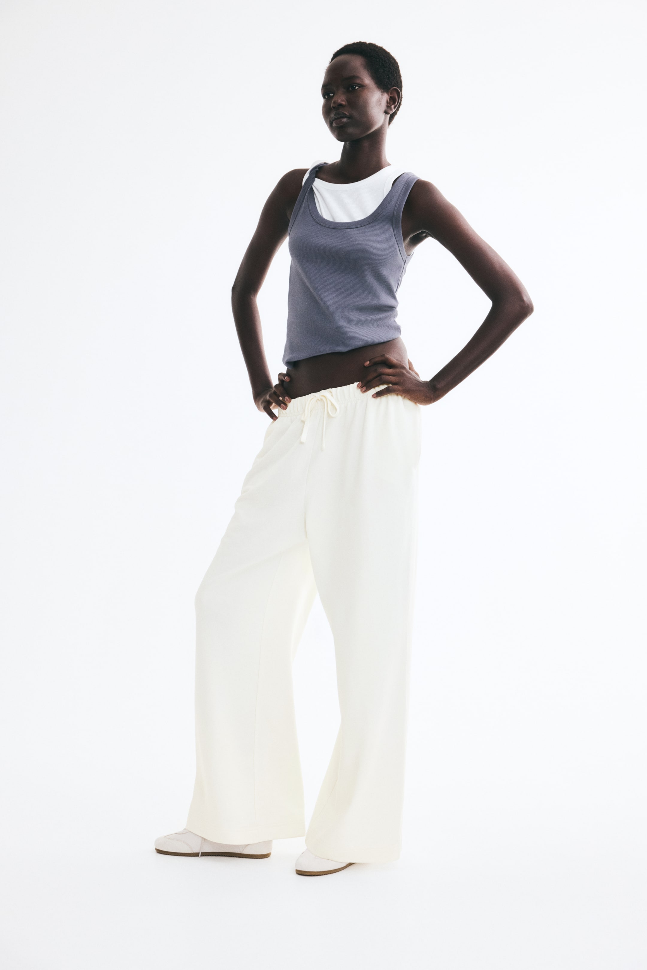 View larger image: Off-white track pants in lightweight sweat fabric are worn by the model, featuring an elasticated waistband with a visible drawstring and straight legs. The pants are styled with layered tank tops and light-colored sneakers.