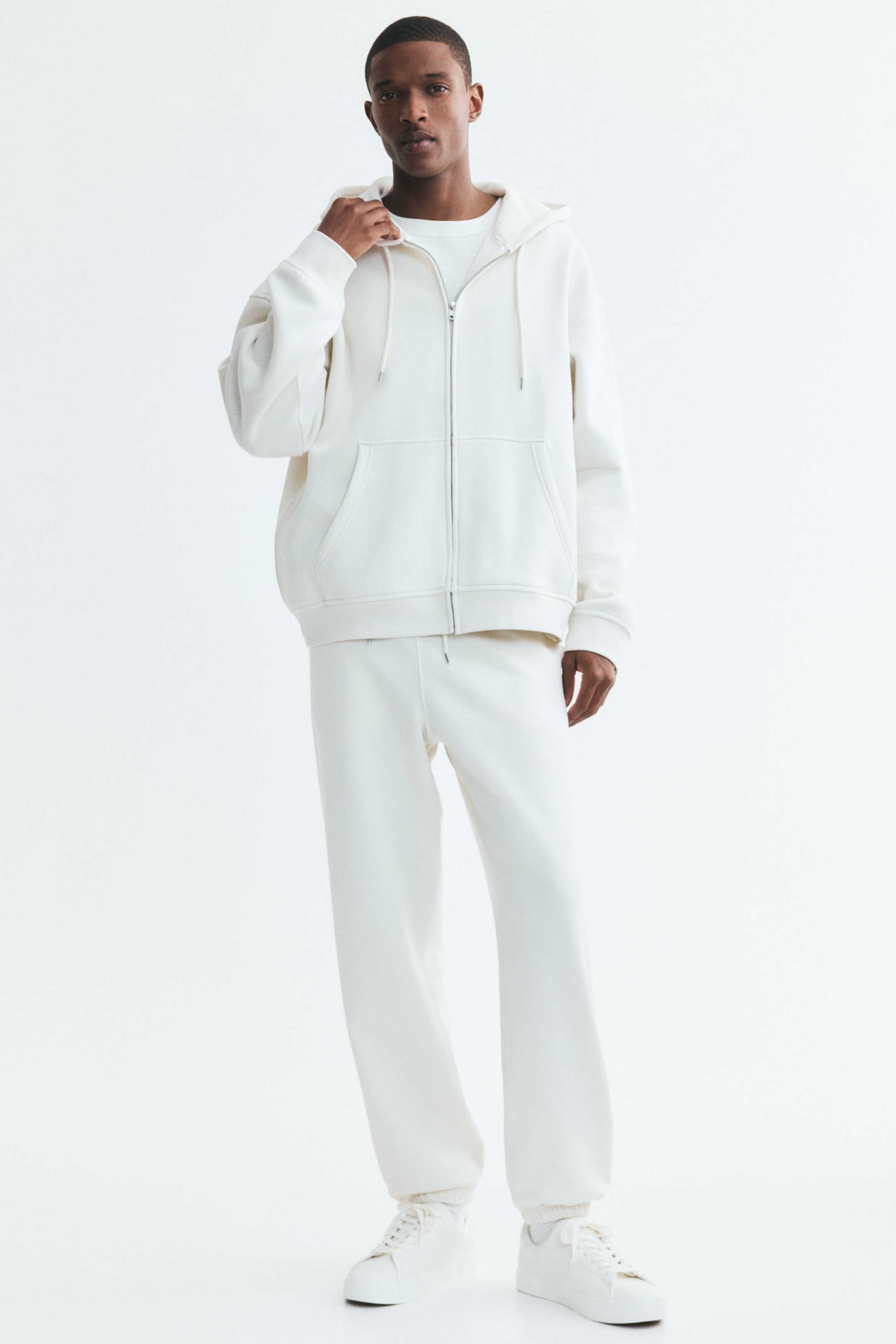 View larger image: The model is sporting cream sweatpants with a relaxed fit, complete with a drawstring waist and ribbed hems. They are styled with a coordinating cream hoodie and white sneakers.