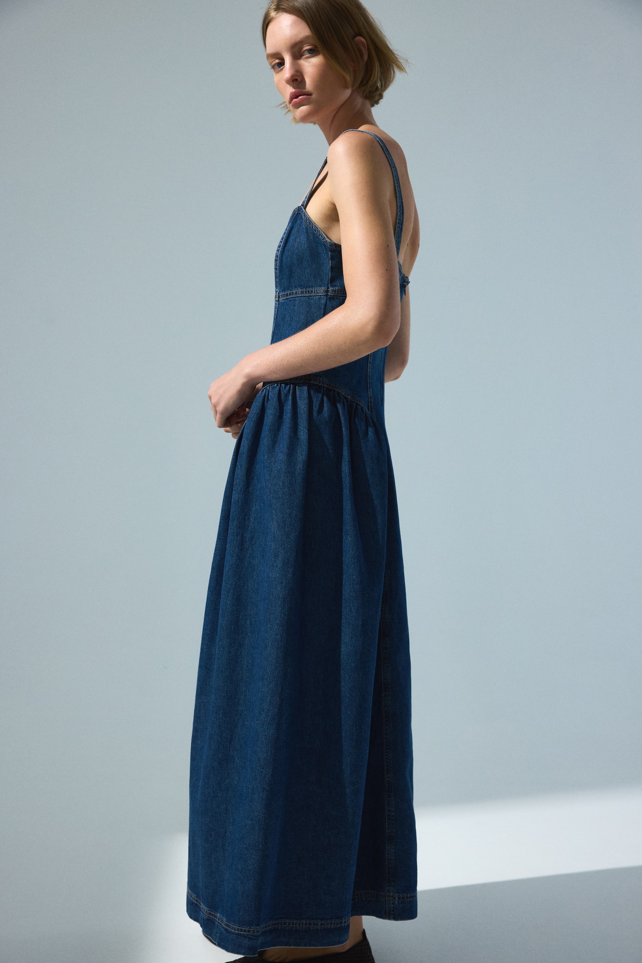View larger image: A dark blue denim dress featuring thin spaghetti straps and a fitted, paneled bodice flows into a wide, gathered skirt. The dress showcases visible contrast stitching detailing its seams and hem, captured from a three-quarter side view.