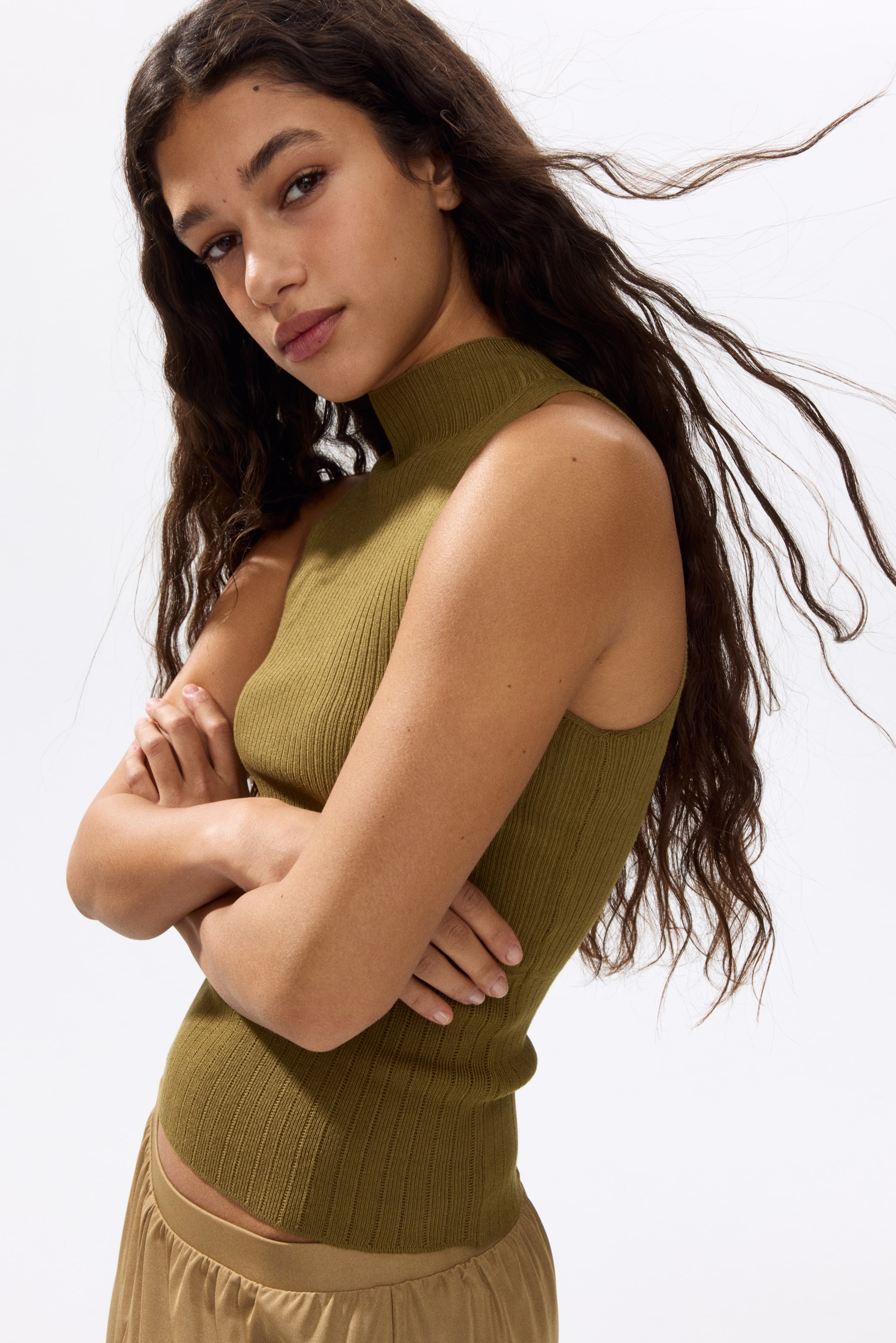 View larger image: An olive green ribbed sleeveless top features a mock neck and a fitted silhouette. Its hem sits just above a light brown waistband.
