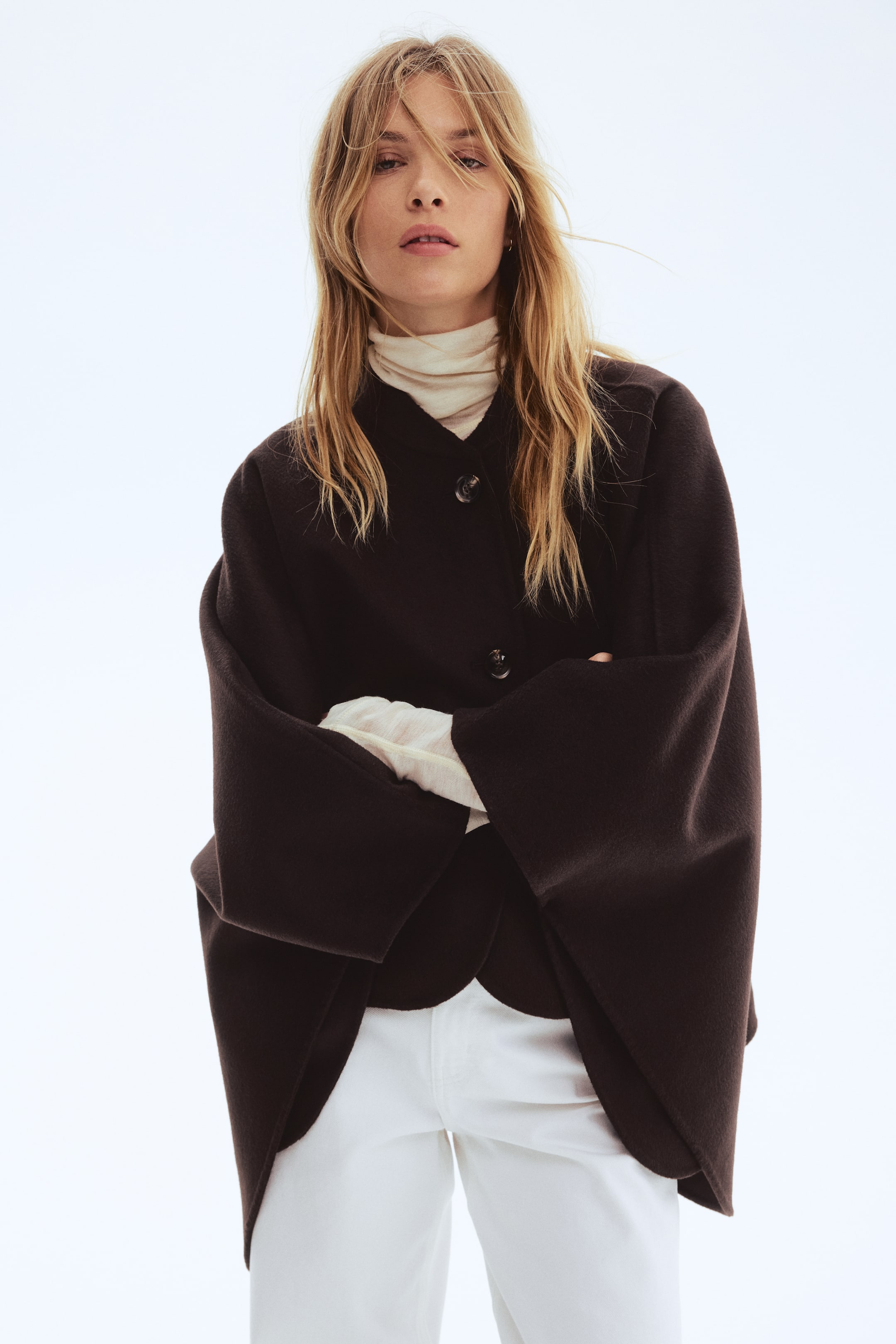 Größeres Bild ansehen: Eine dunkelbraune kurze Jacke mit V-Ausschnitt und einer Dreiknopf-Verschlussleiste. Sie zeichnet sich durch weit geschnittene Ärmel und einen abgerundeten vorderen Saum aus, getragen über einem cremefarbenen Rollkragenpullover und weißen Hosen.