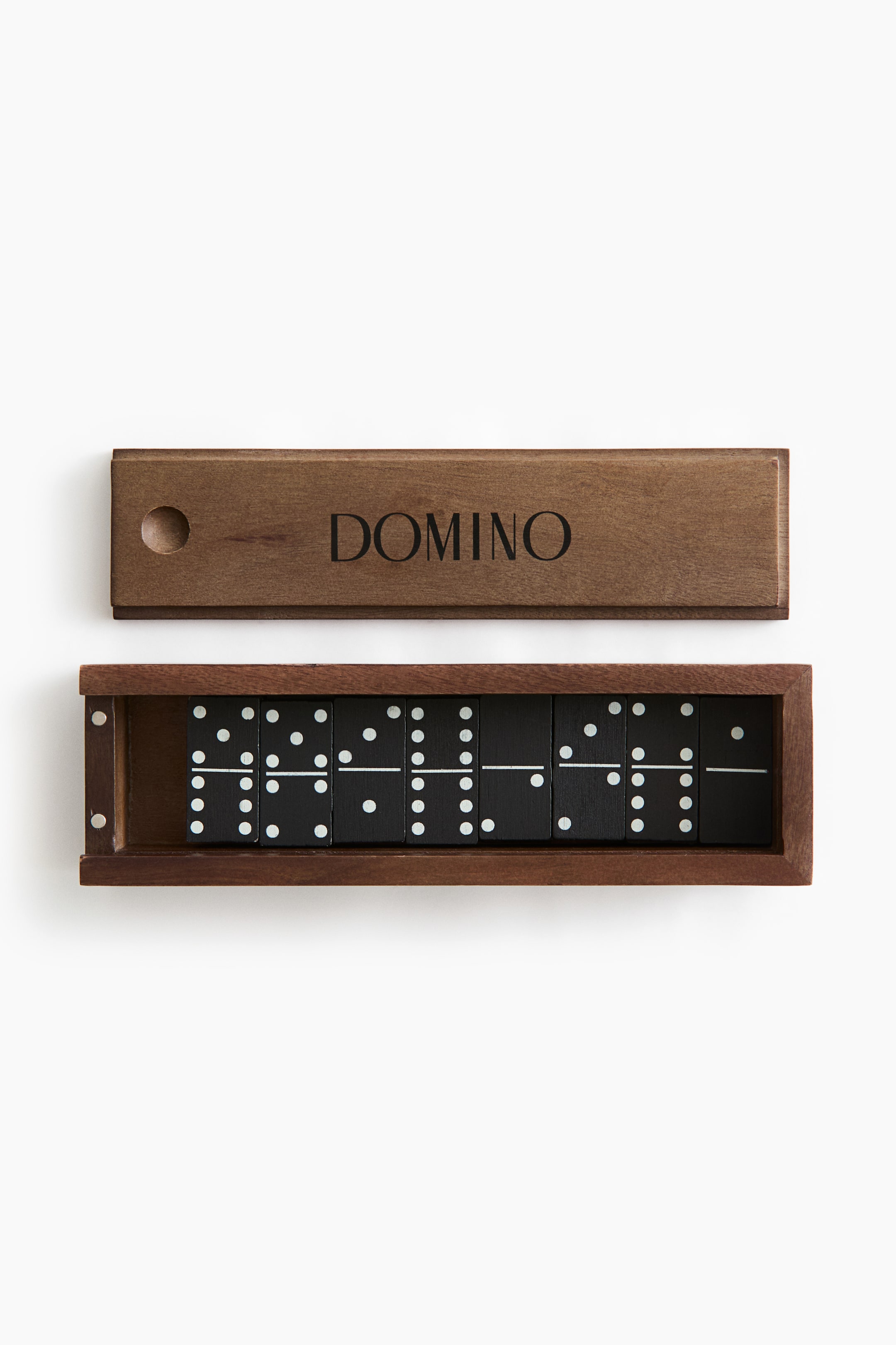 View larger image: A wooden domino set is shown from a top-down perspective, featuring an open wooden box containing black domino tiles with white pips and a matching sliding lid positioned above. The sliding lid displays the word "DOMINO" in a black serif font next to a circular finger indent.