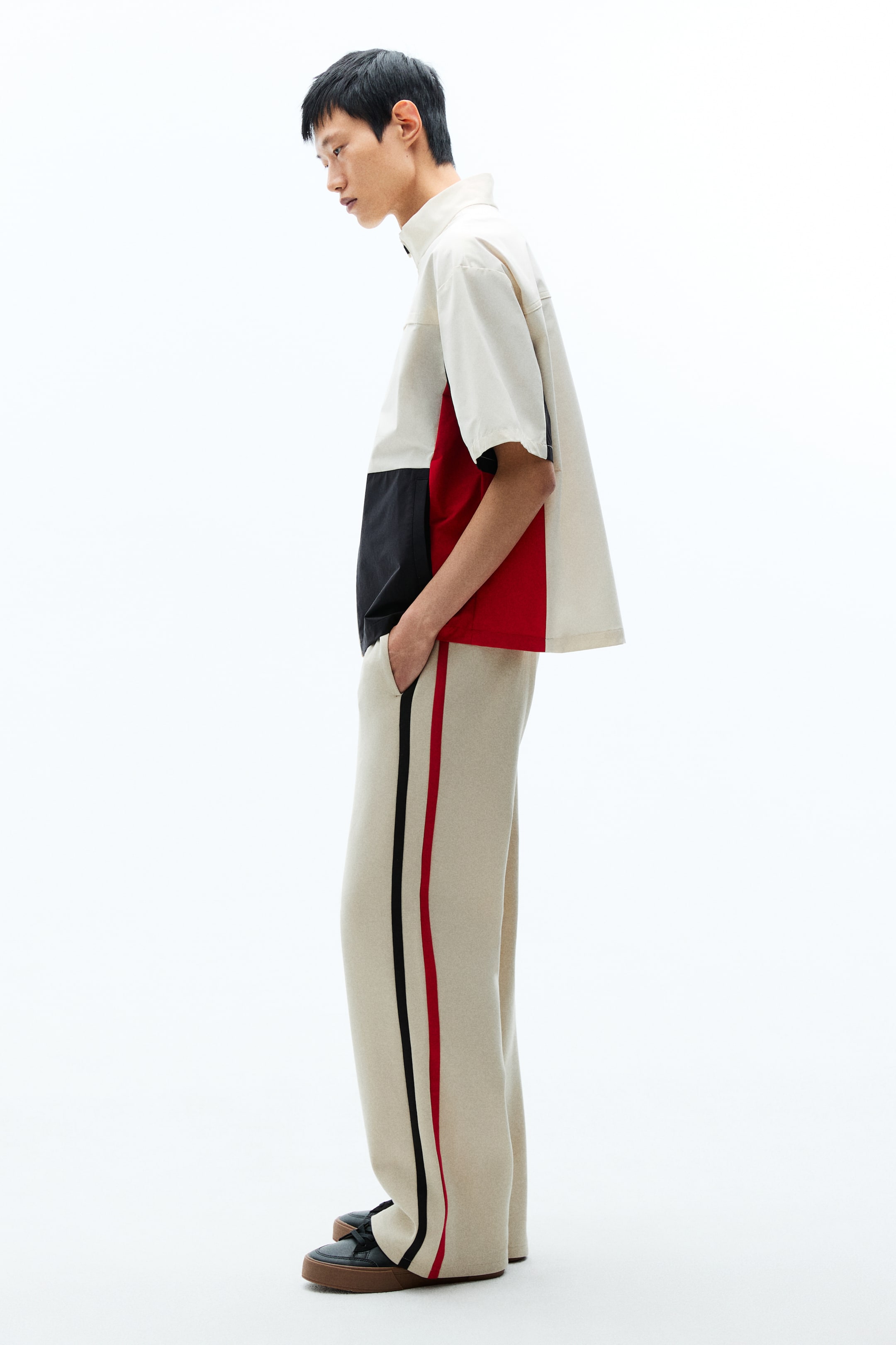 View larger image: The product is a cream, black, and red color-blocked short-sleeved quarter-zip shirt, shown from the side and paired with cream wide-leg pants featuring vertical black and red stripes along each side seam.
