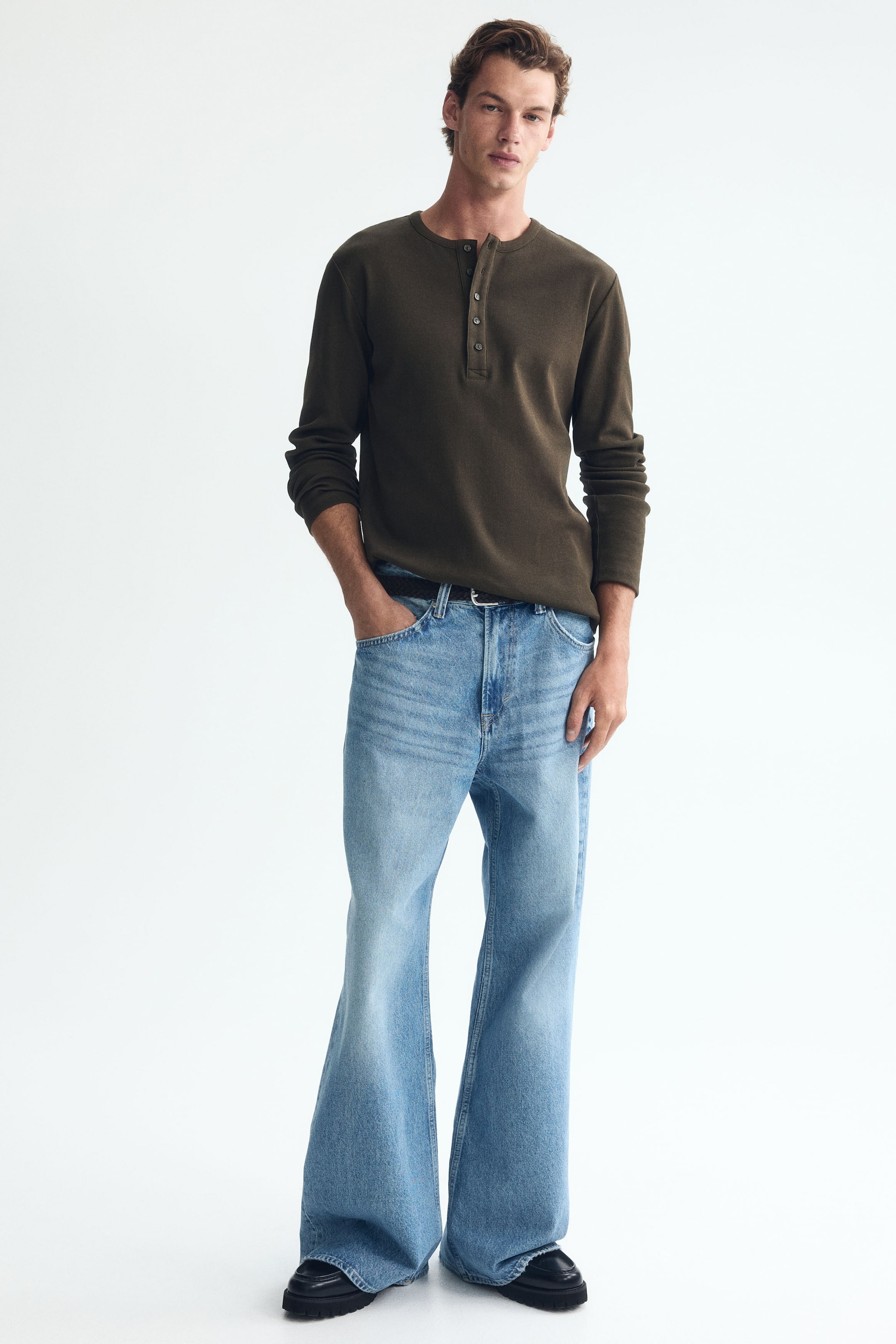 Visa större bild: Femficksjeans i ljusblått, stumt bomullsdenim, med lös passform från sätet till fållen och en lätt utsvängd benform, bärs av modellen. Dessa jeans har normal midja, och en svart bälte pryder midjan medan modellens hand vilar i en framficka.