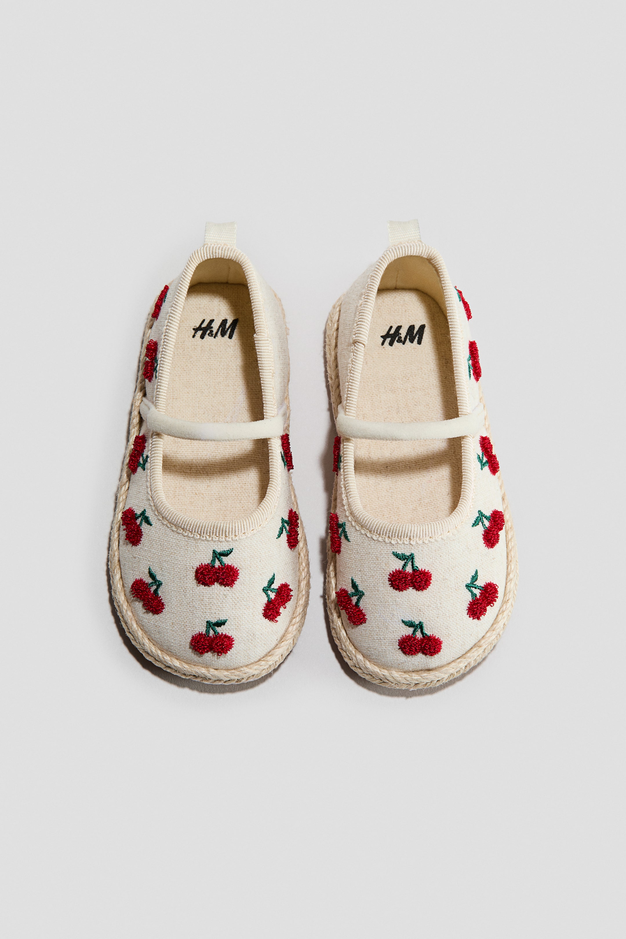 View larger image: A pair of off-white espadrilles is showcased from the front, highlighting rounded toes adorned with red cherry embroidery. An elasticated strap stretches over the foot, while a loop is visible at the back. The soles are encircled by braided jute trim, and the interior reveals a canvas lining.