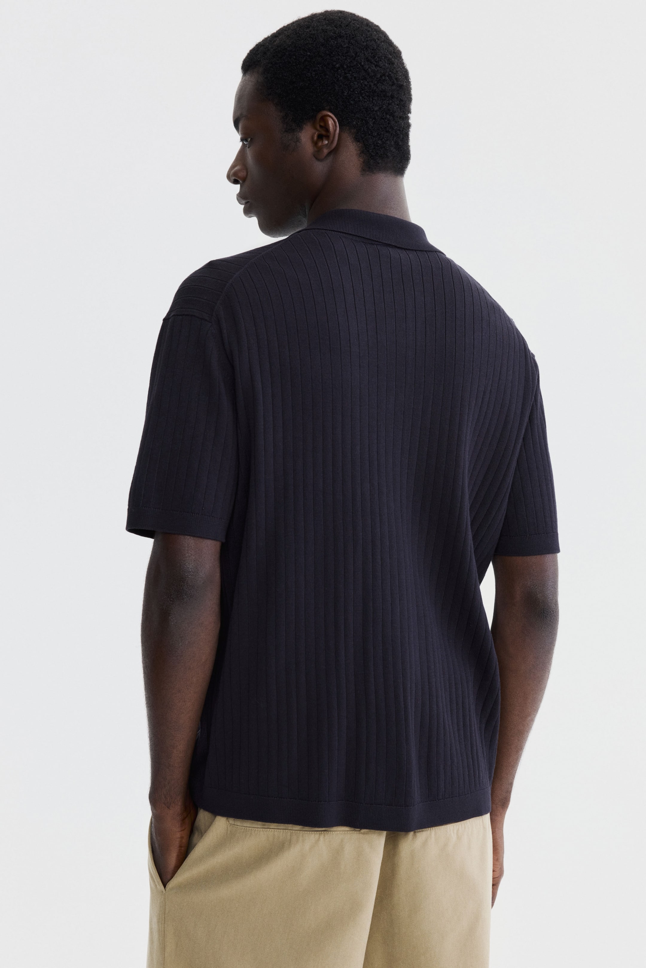 Agrandir l'image: Un polo en maille côtelée bleu foncé est présenté de dos, affichant un col polo classique, des manches courtes avec des poignets côtelés, et un ourlet côtelé, tandis qu'un motif côtelé vertical constant parcourt l'ensemble du corps et des manches.
