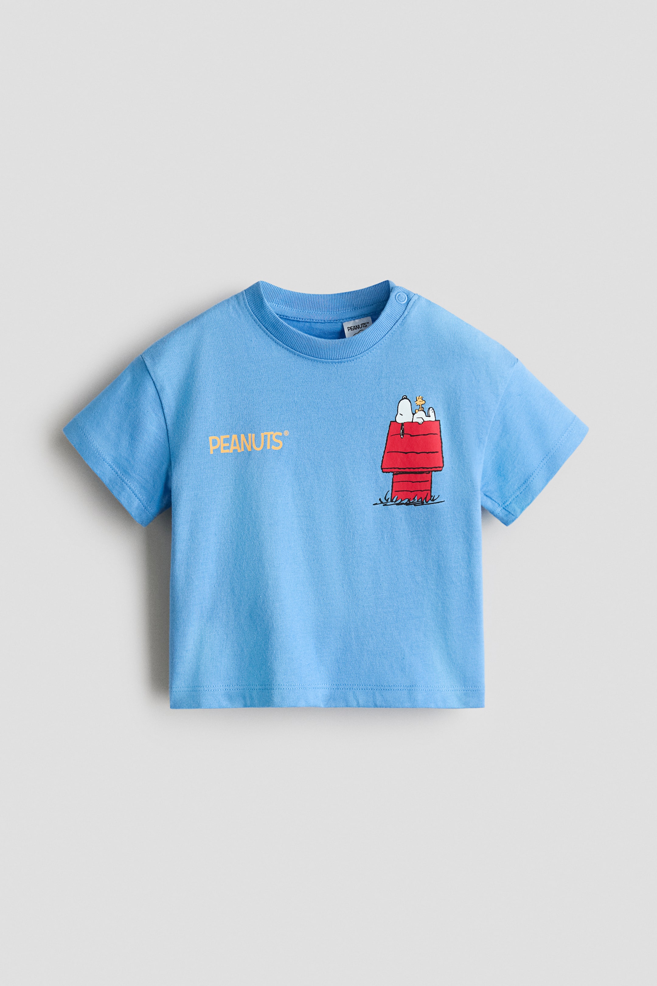 View larger image: A light-blue short-sleeved T‑shirt is shown front-facing, featuring a ribbed crew neck and two snap buttons on the left shoulder. The front displays "PEANUTS" in yellow text on the left chest, alongside an illustration of Snoopy lying on his red doghouse with Woodstock to the right.