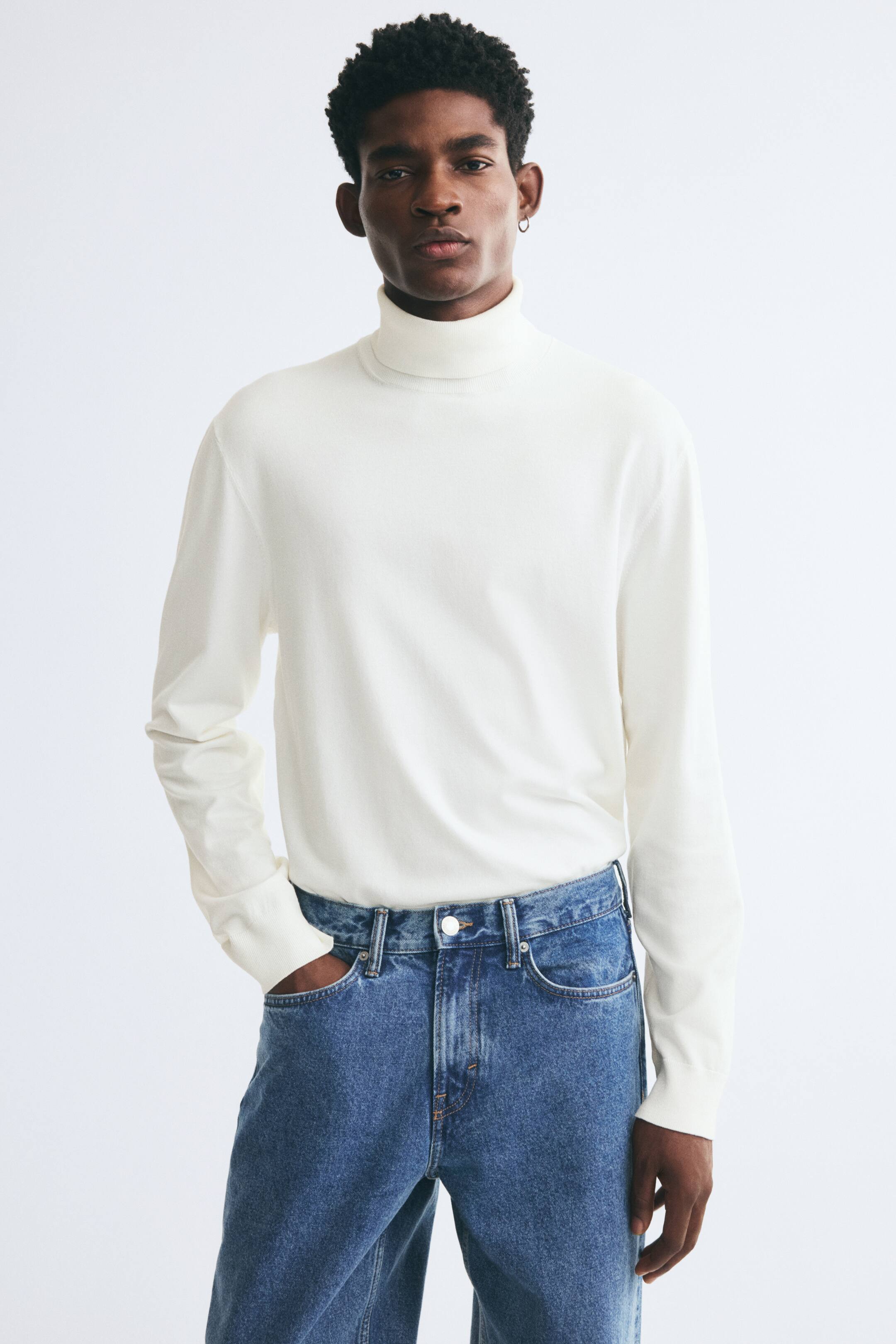 Größeres Bild anzeigen: Das Model trägt einen weißen, fein gestrickten Rollkragenpullover, der in blaue Jeans gesteckt ist. Der Pullover hat eine Regular Fit-Passform mit langen Ärmeln, gerippten Bündchen und einem klassischen, hoch geschnittenen, gerippten Kragen.