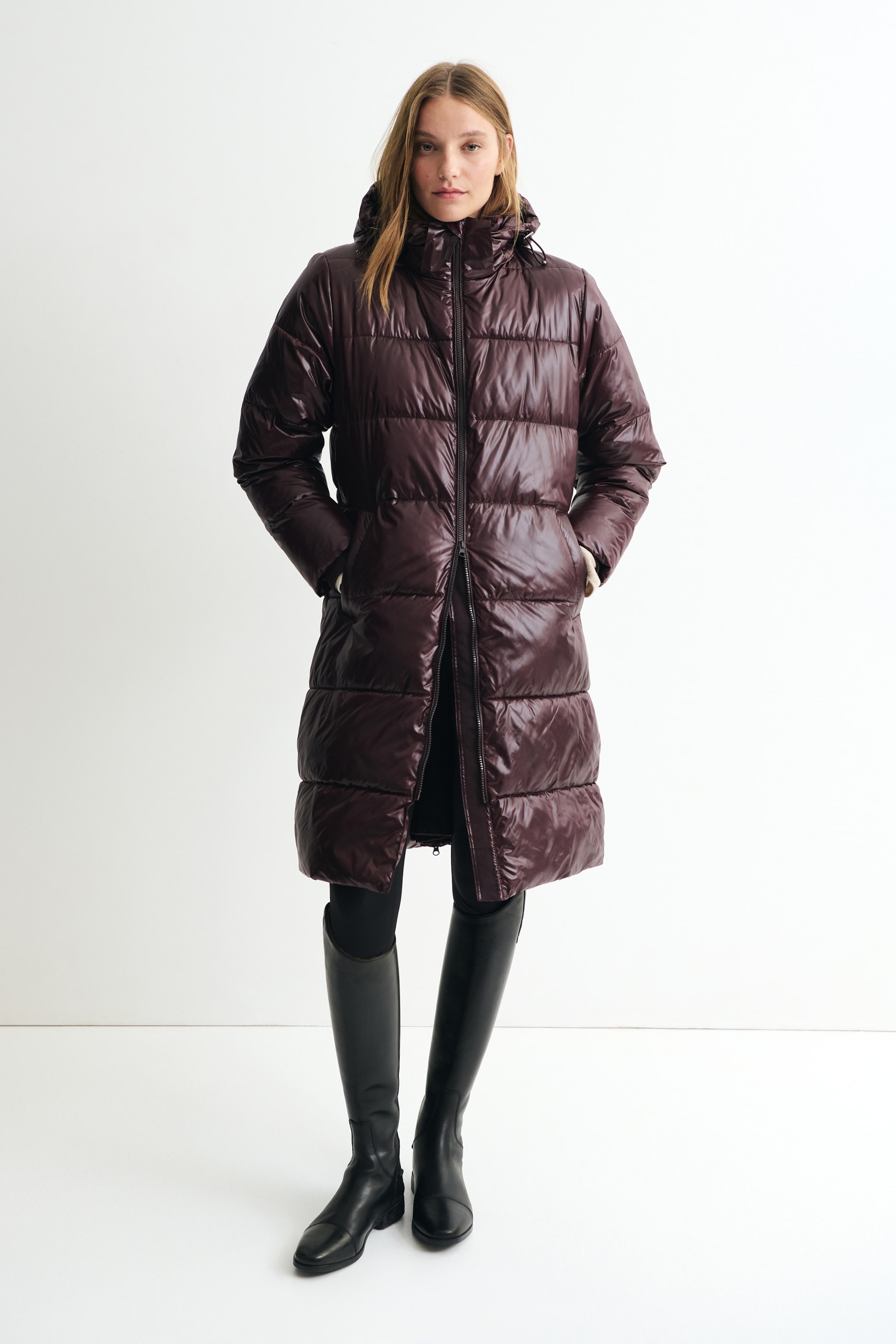 View larger image: A long, glossy dark plum puffer coat is shown from the front, featuring horizontal quilting, a high-neck hood and a front zip fastening. The coat is knee-length with side pockets and is styled with black leggings and knee-high black leather boots.