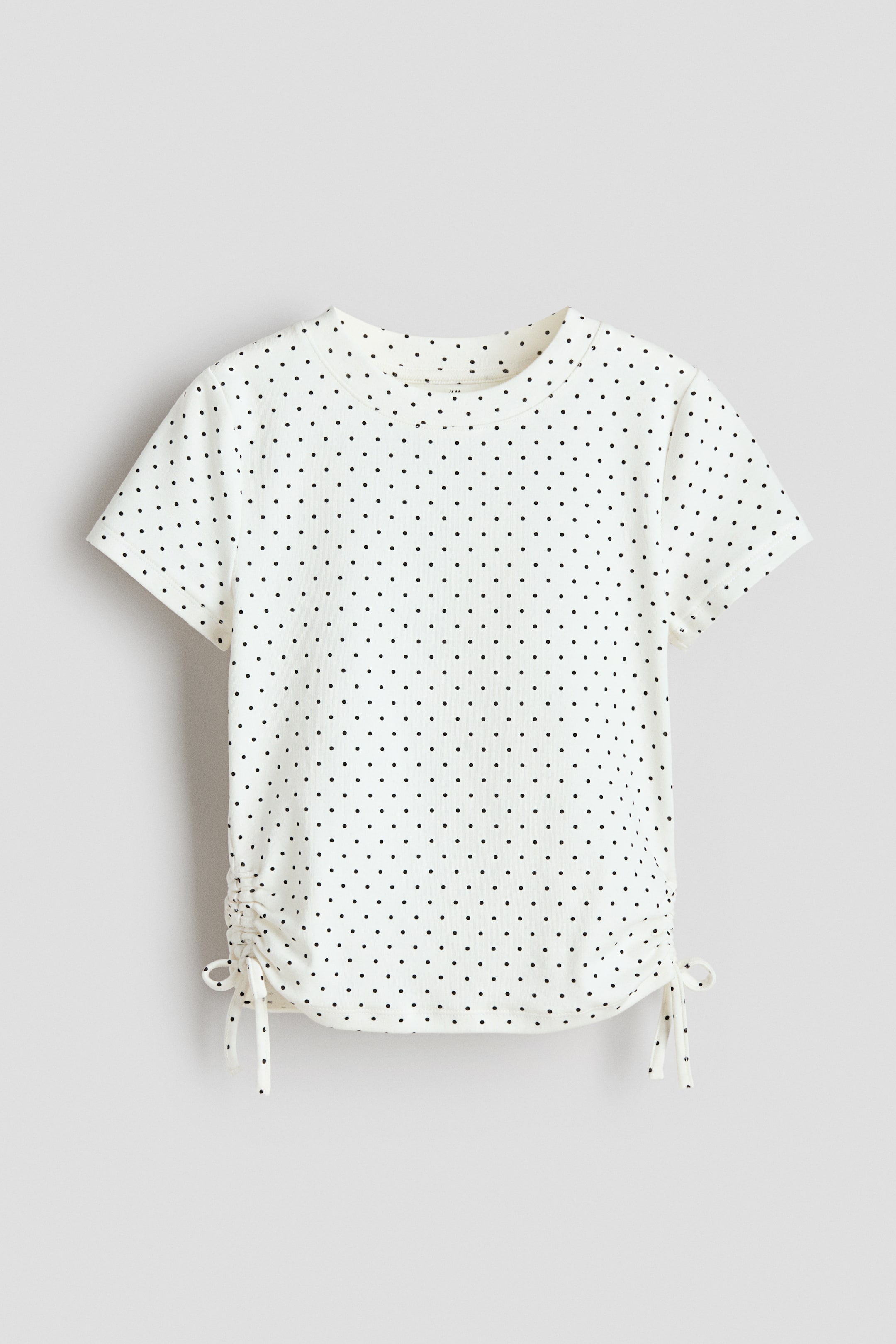 Voir une image plus grande: Un t-shirt blanc à pois noirs présente un col rond, des manches courtes et des cordons de fronces ajustables sur les coutures latérales au bas de l'ourlet, vu de face.