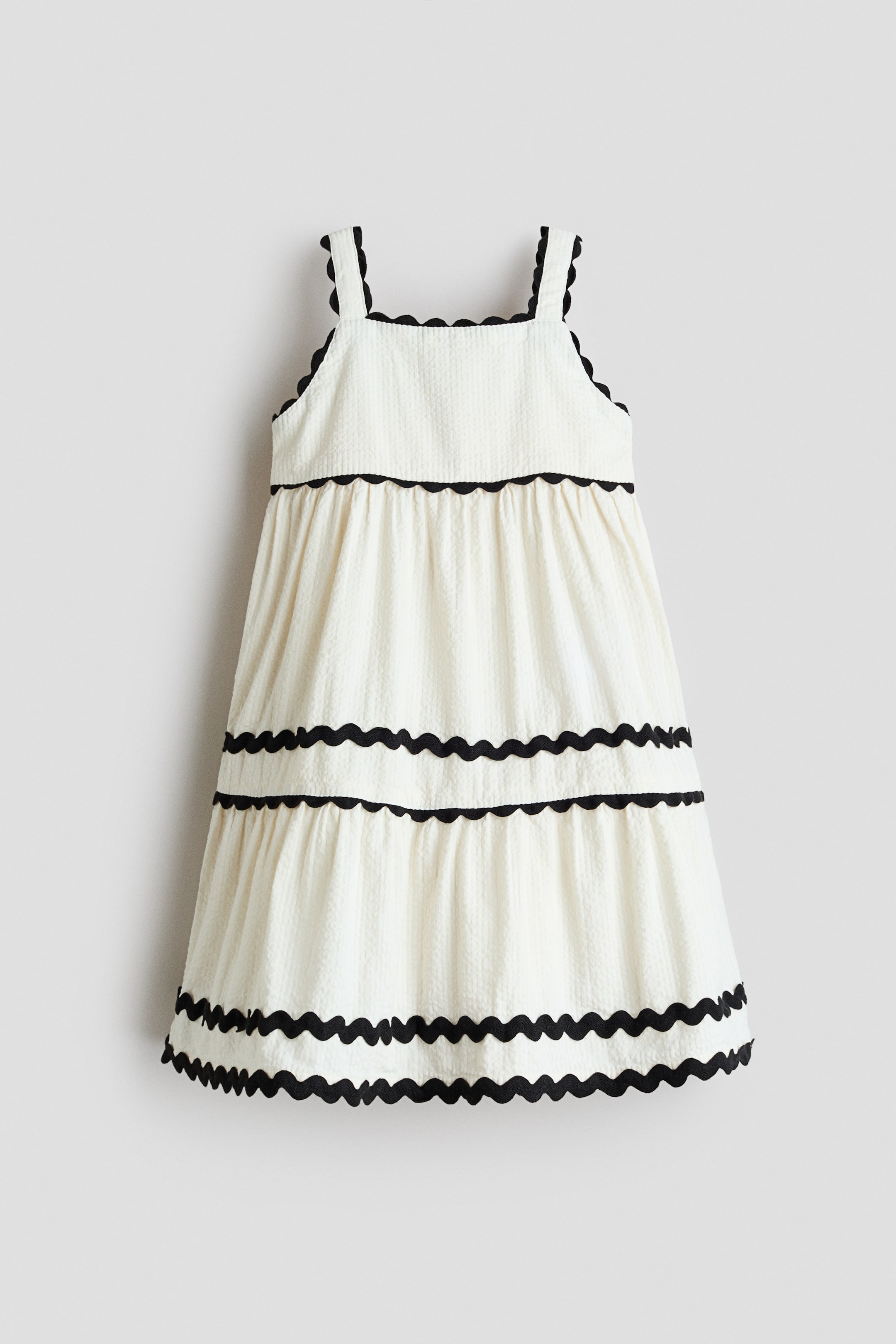 View larger image: A sleeveless dress is displayed from the front, crafted from cream-coloured seersucker fabric. It boasts a square neckline and wide straps, all accented with black scalloped trim, and is tiered with matching black scalloped trim separating each section, creating a flared skirt with gathers for added volume.
