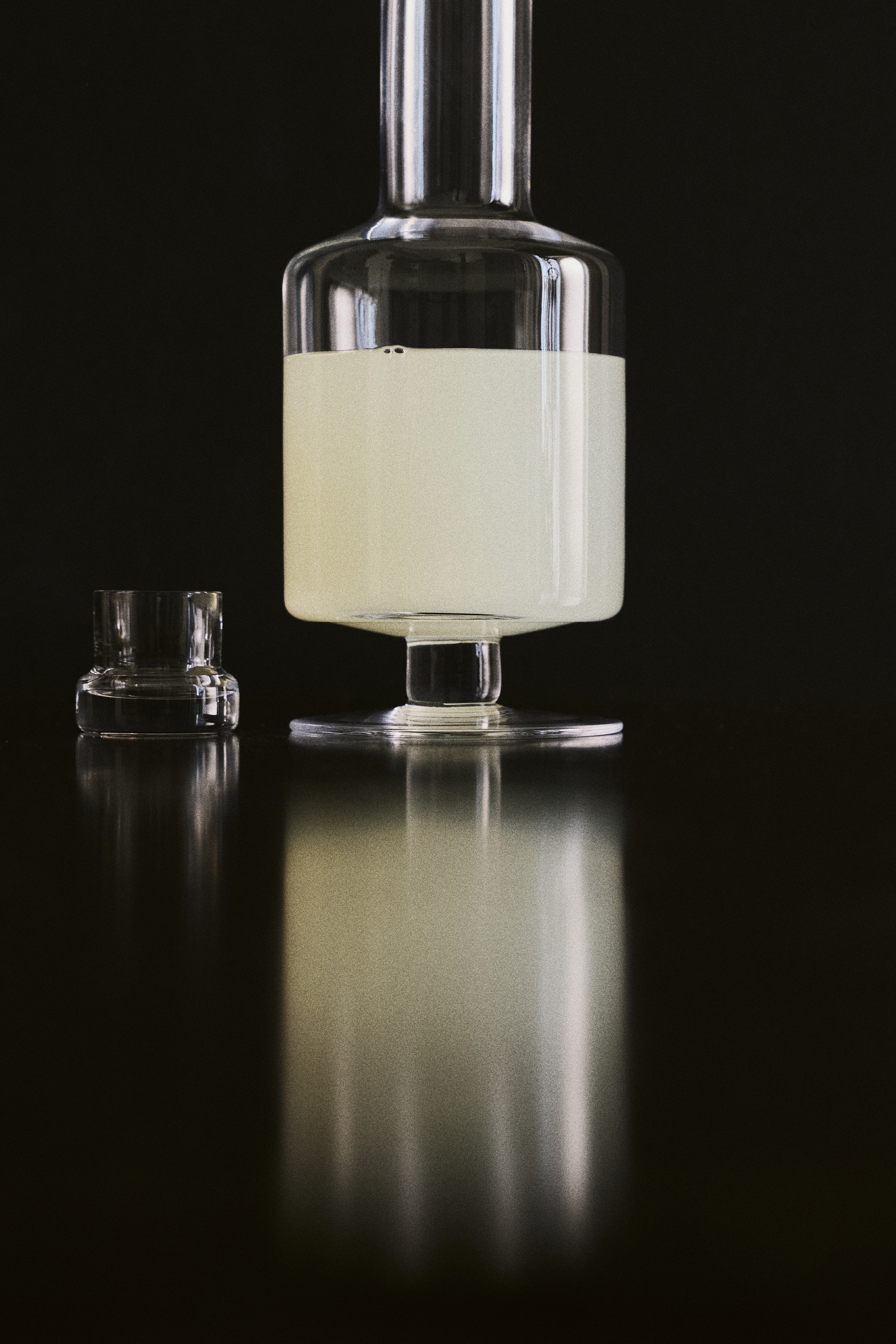 Un decantador de vino con base, visto de frente, presenta una construcción de vidrio transparente soplado a mano con bordes suavemente redondeados. Cuenta con un cuello estrecho, con un tapón de vidrio a juego en su interior, y una base ancha y plana.