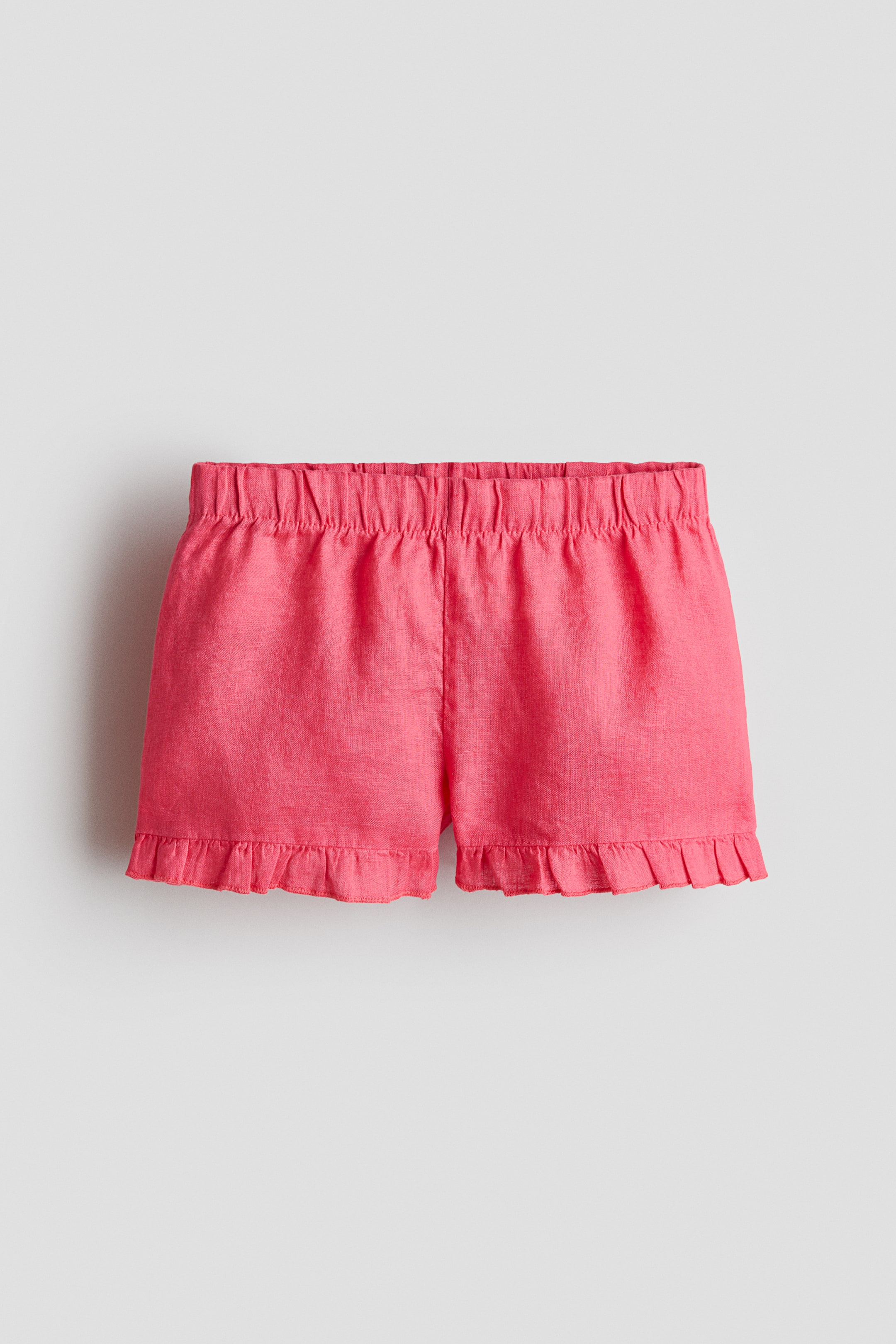 View larger image: Pink linen shorts are shown from the front, featuring an elasticated, gathered waist and delicate frill trims along the hems.