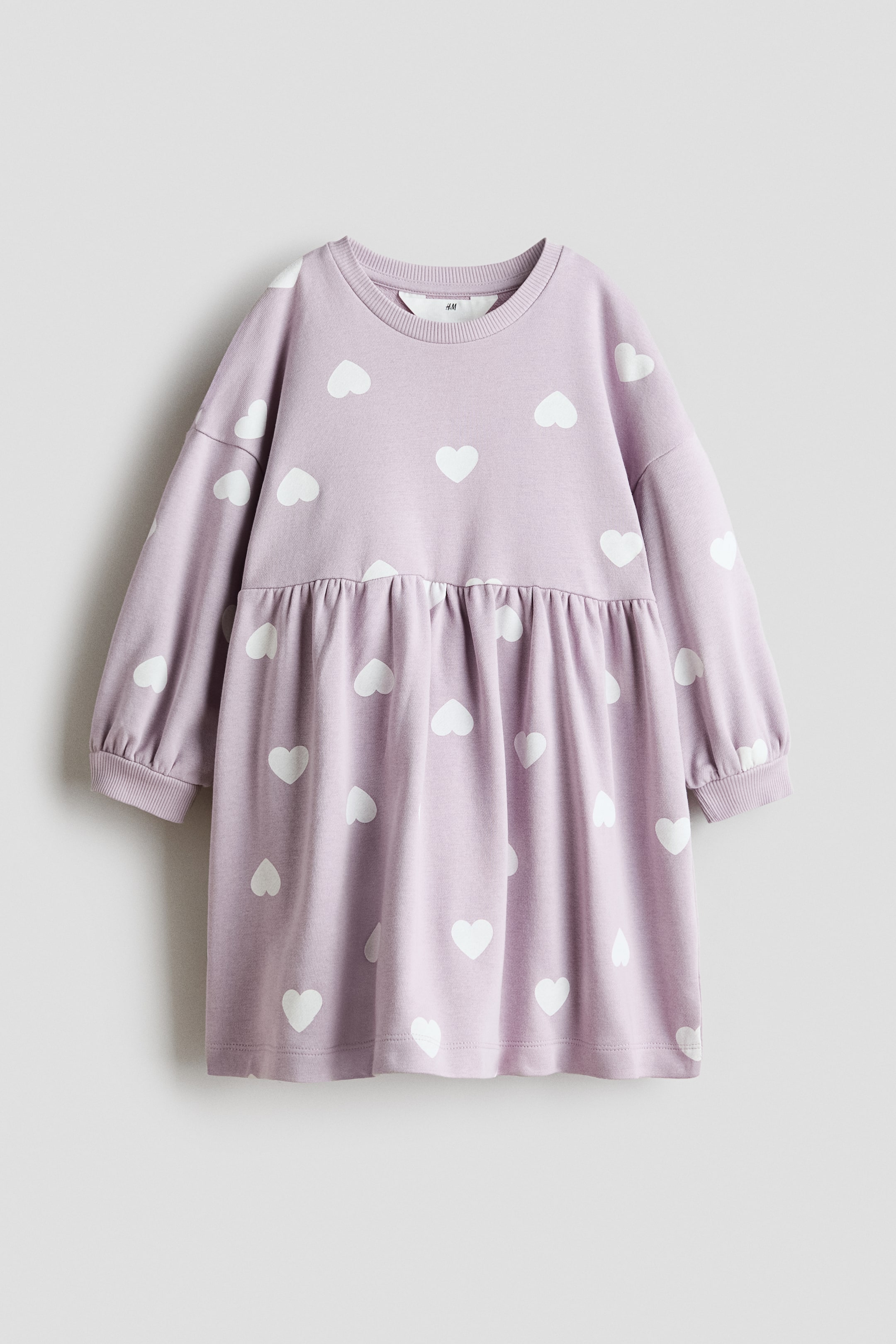 Ingrandisci l'immagine: Vista frontale di un abito oversize in morbido tessuto felpa color lilla chiaro con un motivo di cuori bianchi sparsi. Presenta un girocollo a coste, maniche lunghe leggermente più ampie con polsini a coste e una cucitura arricciata sui fianchi che aggiunge ampiezza all'orlo.
