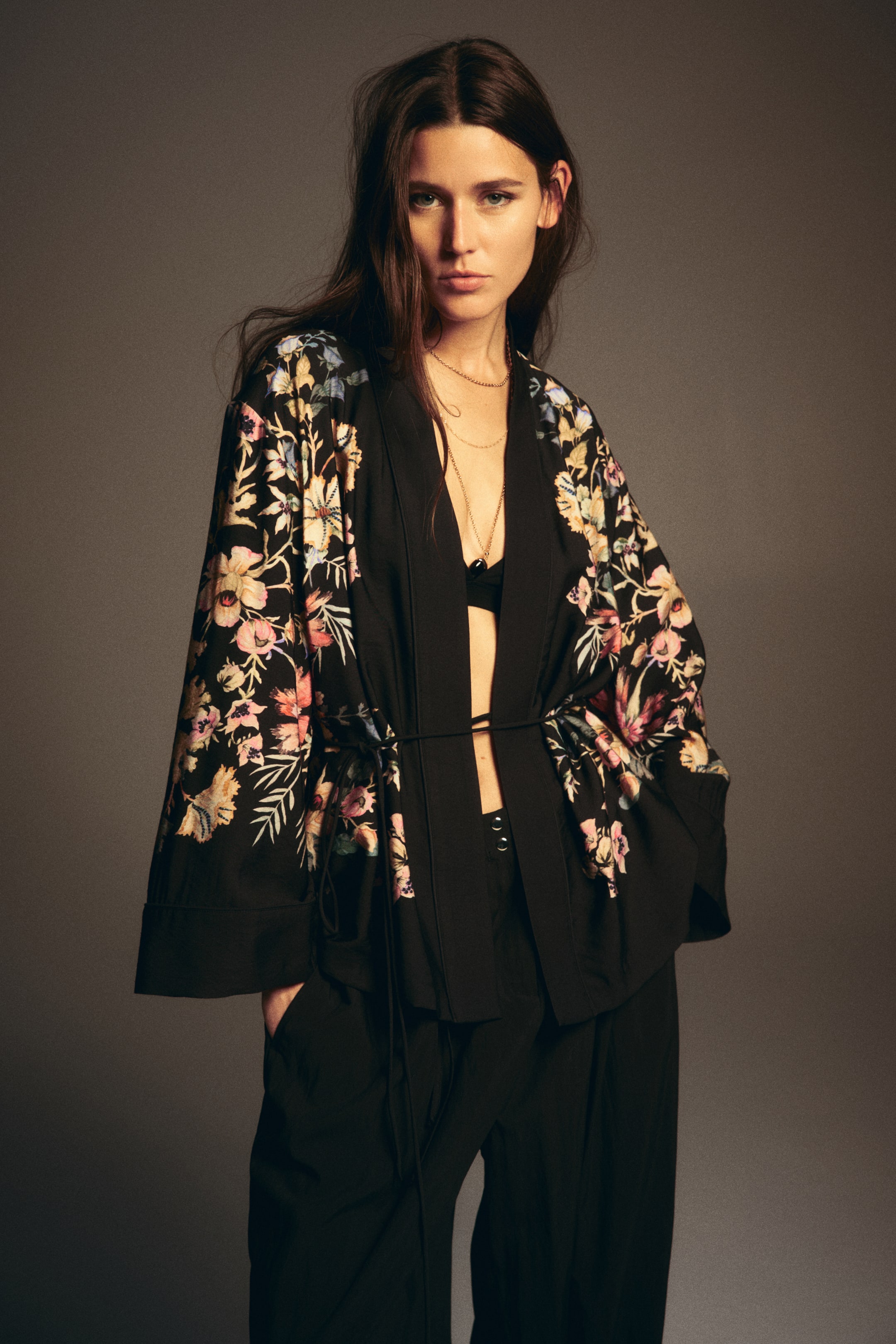 View larger image: A black kimono-style jacket is displaying a vibrant floral print in shades of pink, yellow, blue, and green across its wide sleeves and upper body. It is featuring an open front, cinched at the waist by a tie closure, revealing a black bralette underneath.