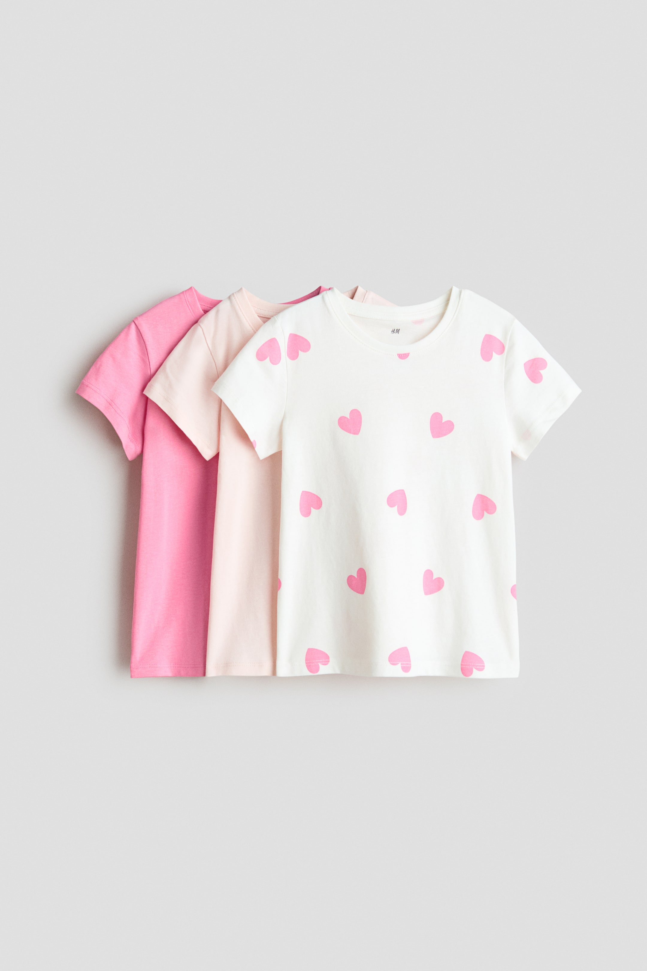 Ver imagen más grande: Tres camisetas de suave punto de algodón presentan un escote redondo y un dobladillo recto, vistas desde el frente. Una camiseta es de un color sólido rosa intenso, otra es de un color sólido rosa claro, y la camiseta delantera muestra un patrón de corazones rosas dispersos sobre una base blanco roto.