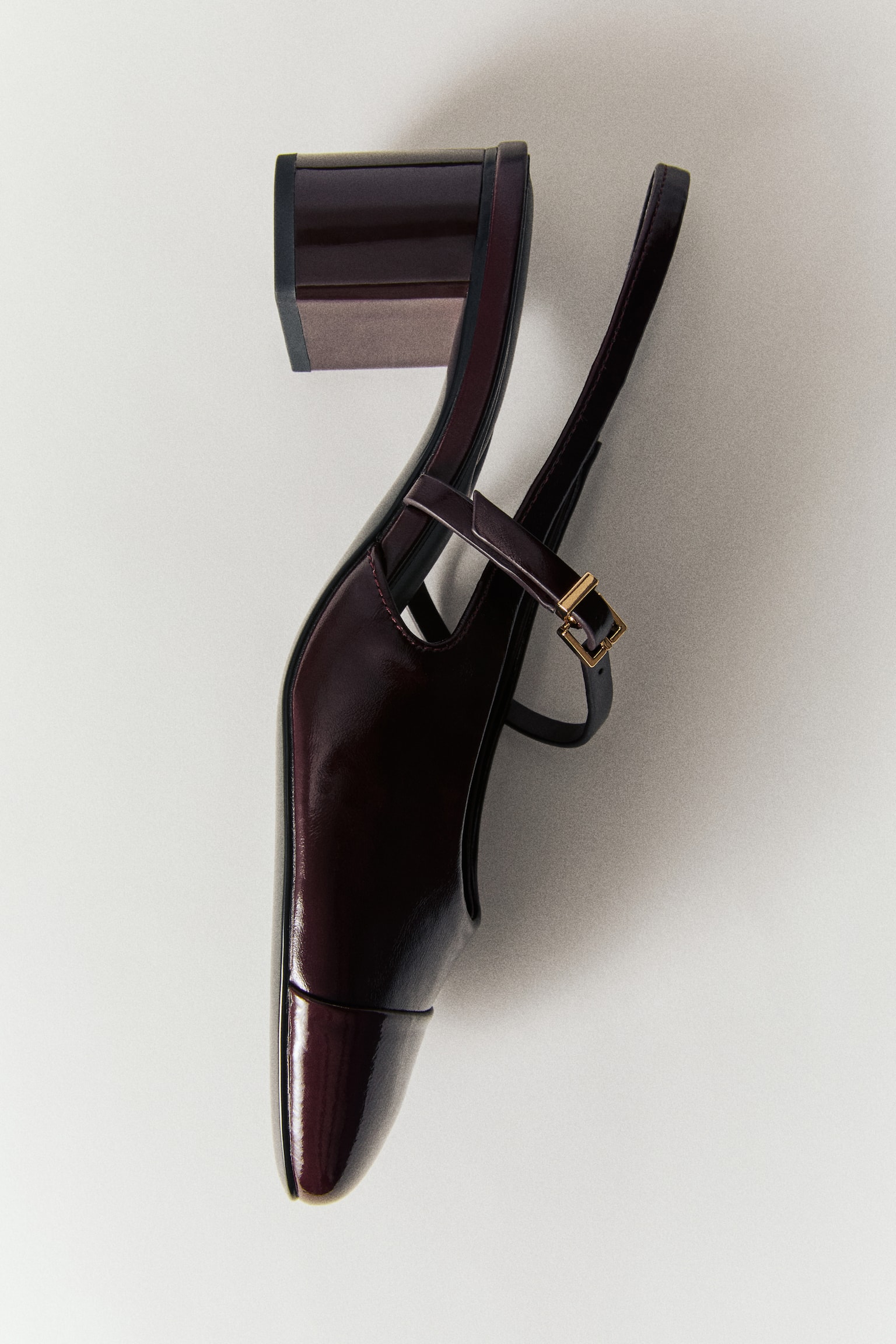 A dark burgundy patent leather Mary Jane shoe is shown in a close-up, front-facing view, highlighting its glossy, high-shine finish. It features a structured square cap toe and a slim, adjustable instep strap with a small, gold-tone rectangular buckle. - 1