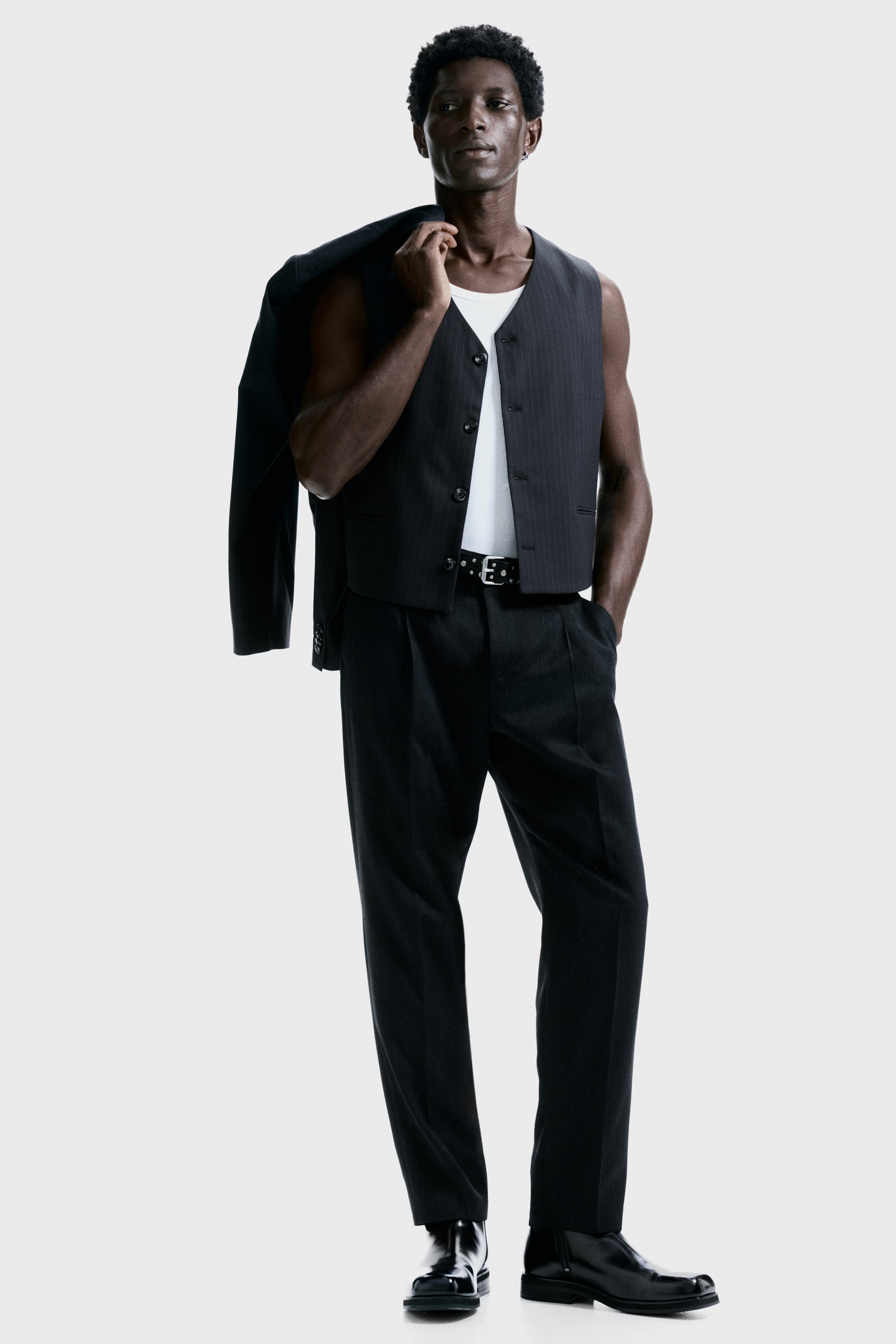 View larger image: A dark pinstriped suit is being presented from a slightly angled front view, featuring a vest with a button-front closure and cropped hem, worn over a white tank top. Matching pleated trousers are being styled with a black studded belt, while a coordinating dark jacket is being draped over the left shoulder.