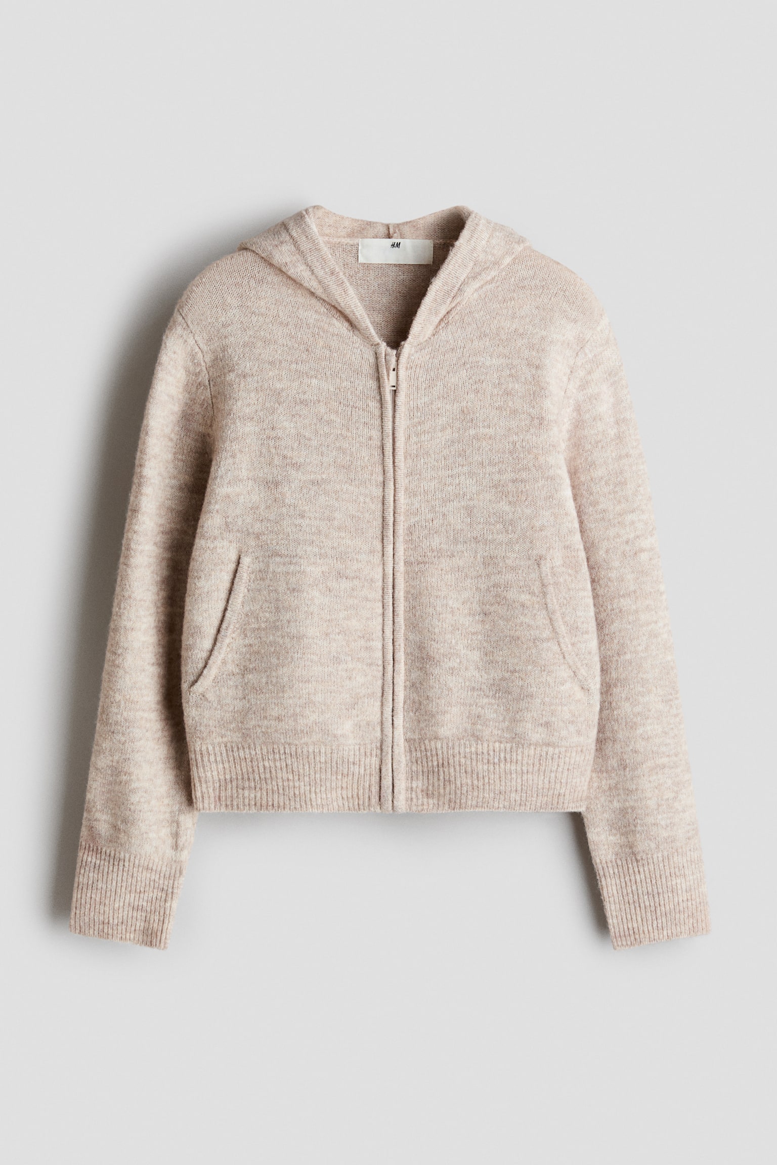 Een lichtbeige gebreid hoodievest met rits wordt van voren getoond en heeft een zachte, licht geborstelde textuur. Het kledingstuk is voorzien van een centrale ritssluiting, schuine steekzakken aan de voorkant, een capuchon met fijne ribboorden en brede ribboorden aan de mouwboorden en zoom.