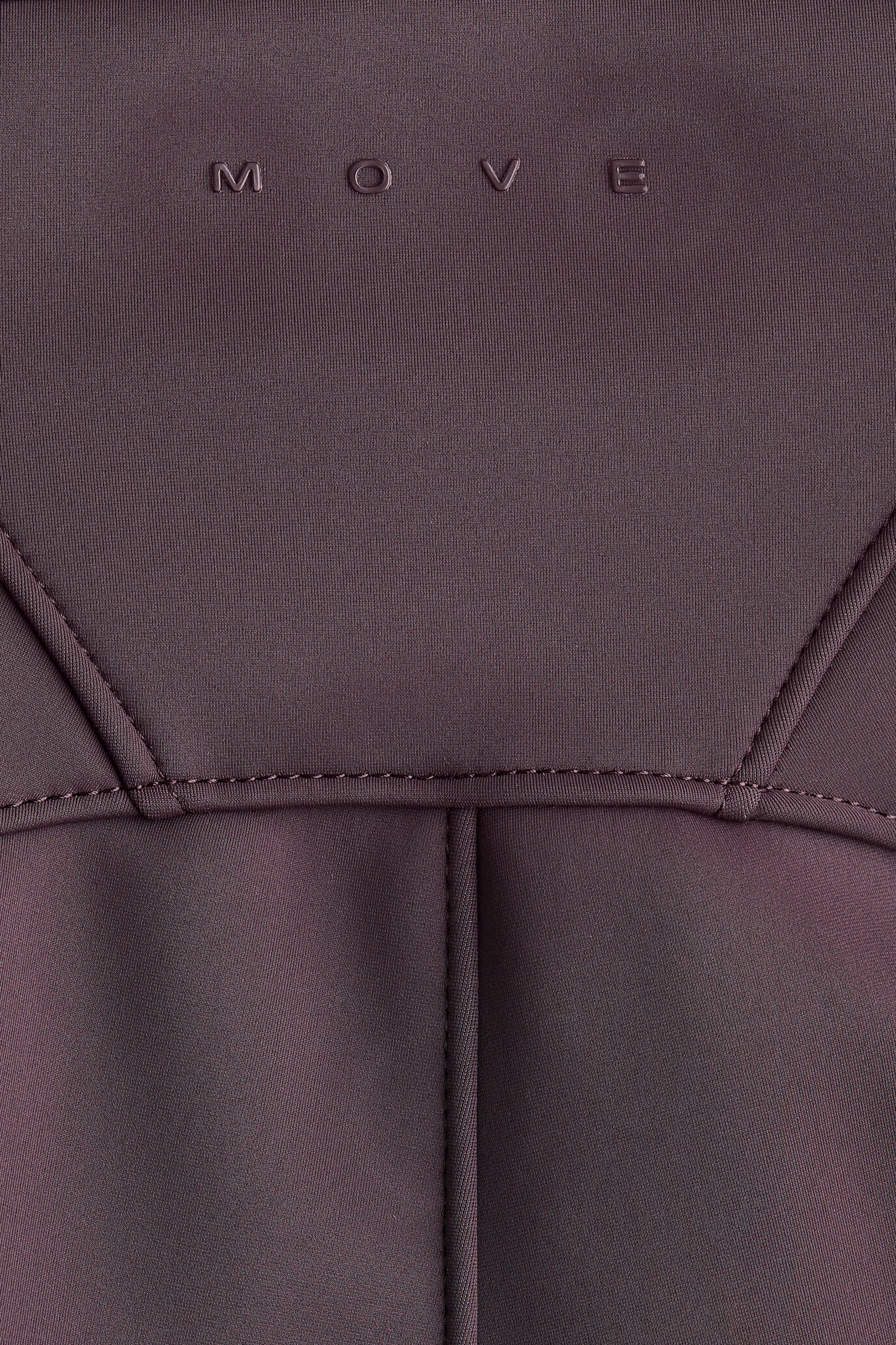 Agrandir l'image: Un gros plan de pantalons de ski prune foncé montre la section supérieure avant, avec un tissu fonctionnel texturé et le lettrage en relief 'MOVE' en haut. Un design structuré est visible avec une couture horizontale et des coutures diagonales s'étendant vers le haut, et des plis verticaux subtils apparaissent sous la couture horizontale.
