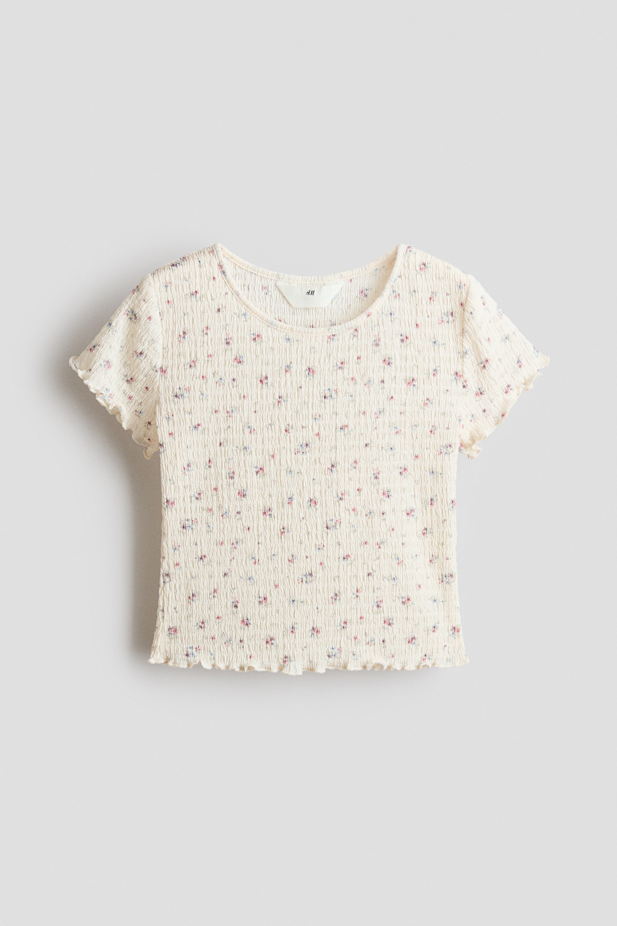 View larger image: A cream short-sleeved top is shown from the front, displaying a shirred texture throughout. It features a small floral print in pink, blue, and purple, a round neckline, and ruffled edges on the sleeves and hem.
