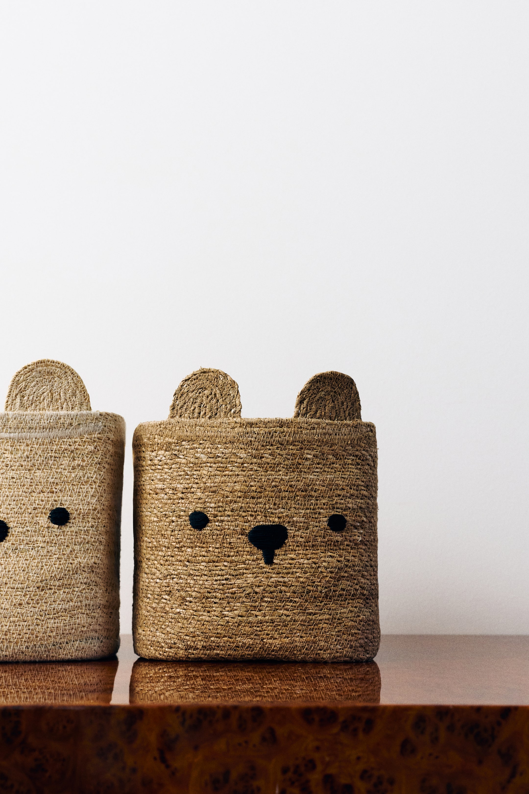View larger image: The Bear Storage Basket, crafted from woven natural fibres, is facing front and features two upright rounded ears along the top rim with black embroidered eyes and a nose on the front surface. A second, lighter-coloured basket of the same design is partially visible on the left.