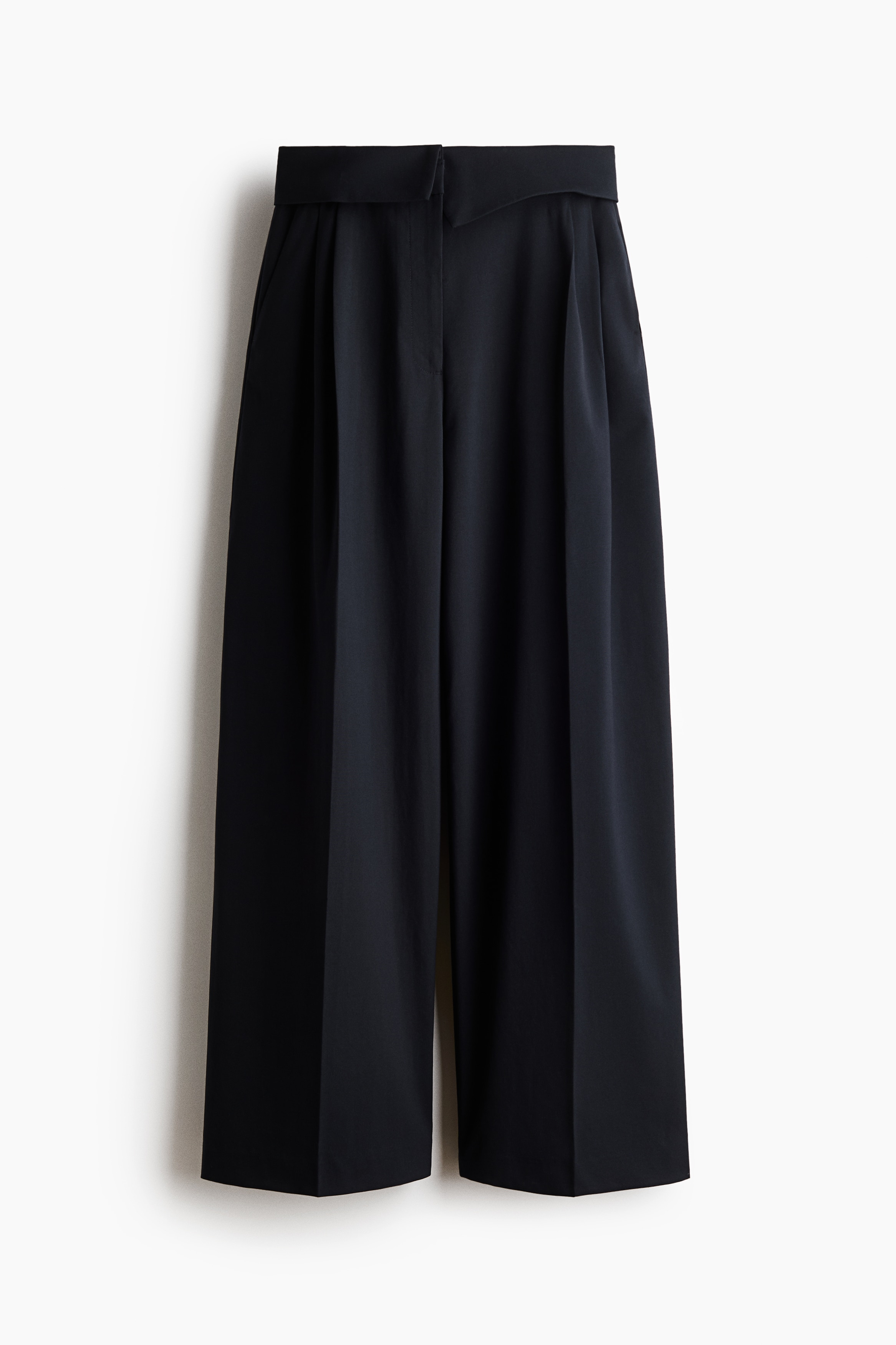 The model is wearing loose-fitting dark woven trousers, featuring a high waist with a wide, overlapping foldover waistband. The trousers have wide legs with front pleats and creases, styled with a dark crew-neck jumper that is partially tucked in to showcase the waistband detail.