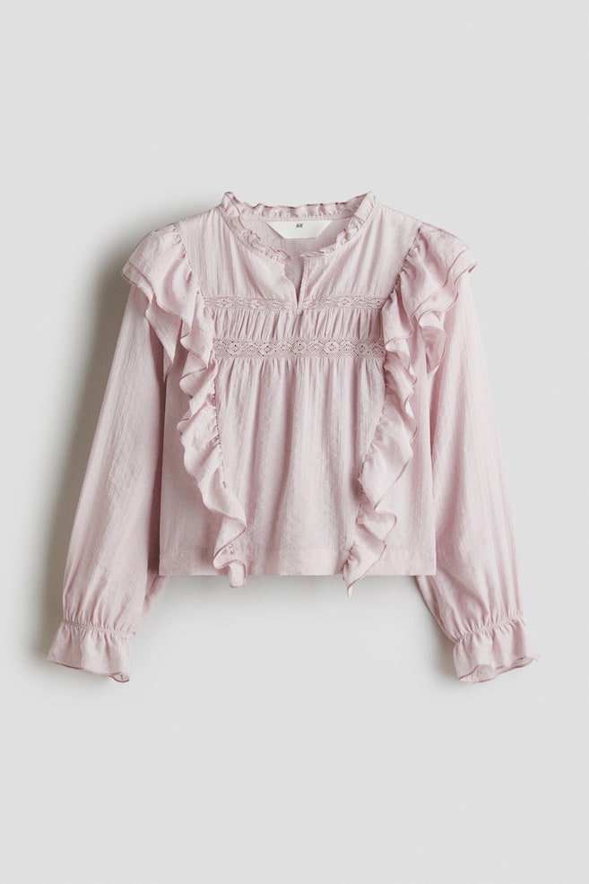 Una blusa rosa chiaro realizzata in un tessuto strutturato mostra la sua vista frontale, caratterizzata da un colletto arricciato a collo alto con uno spacco a V e inserti in pizzo orizzontali sul petto con arricciature sotto. Grandi volant verticali incorniciano il corpetto anteriore e le maniche lunghe terminano con polsini elasticizzati. - 1