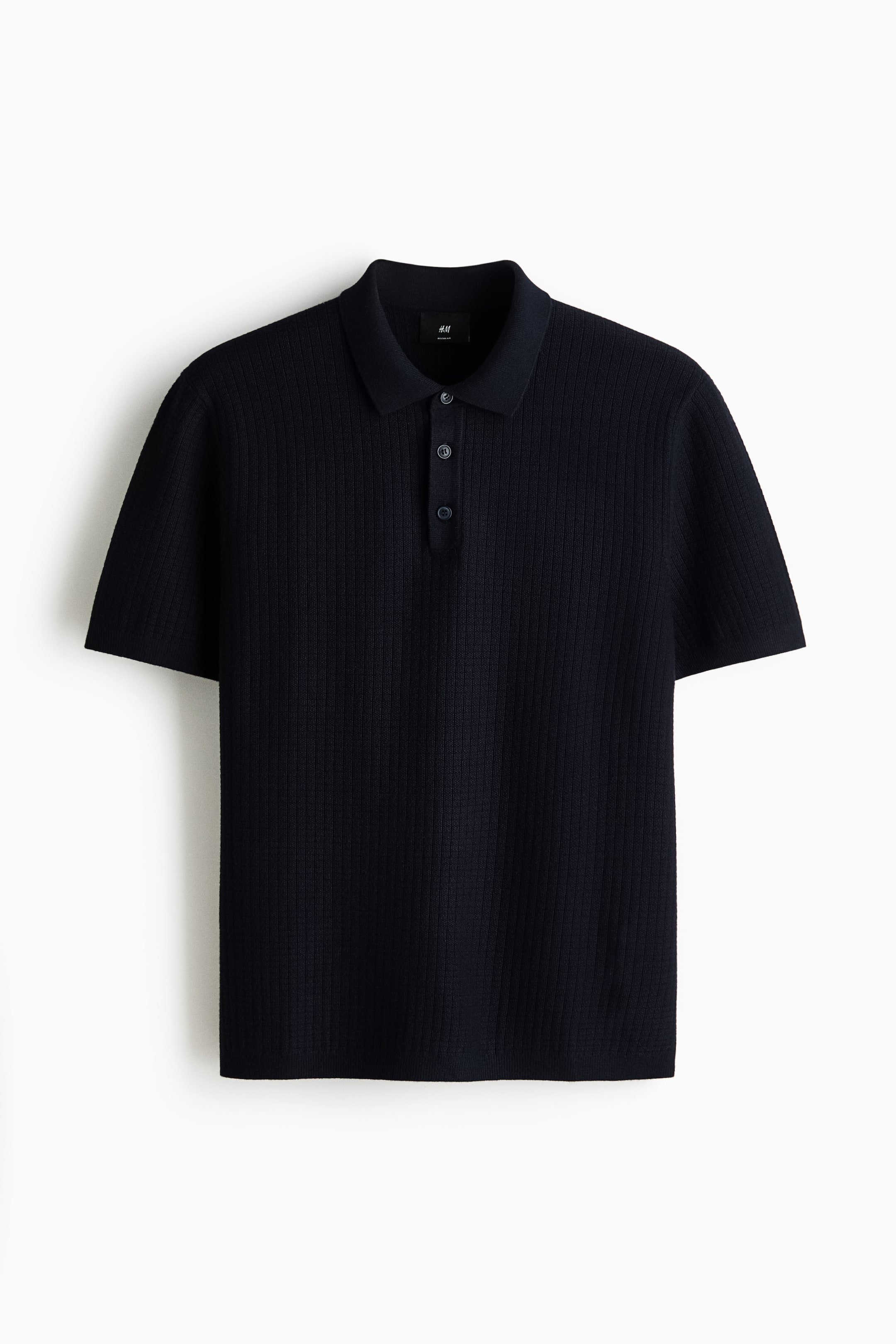 Ver imagen más grande: Una camisa polo azul oscuro está de frente, mostrando una textura de punto acanalado. Está diseñada con un cuello, una tapeta de tres botones, mangas cortas, puños acanalados y un dobladillo acanalado.