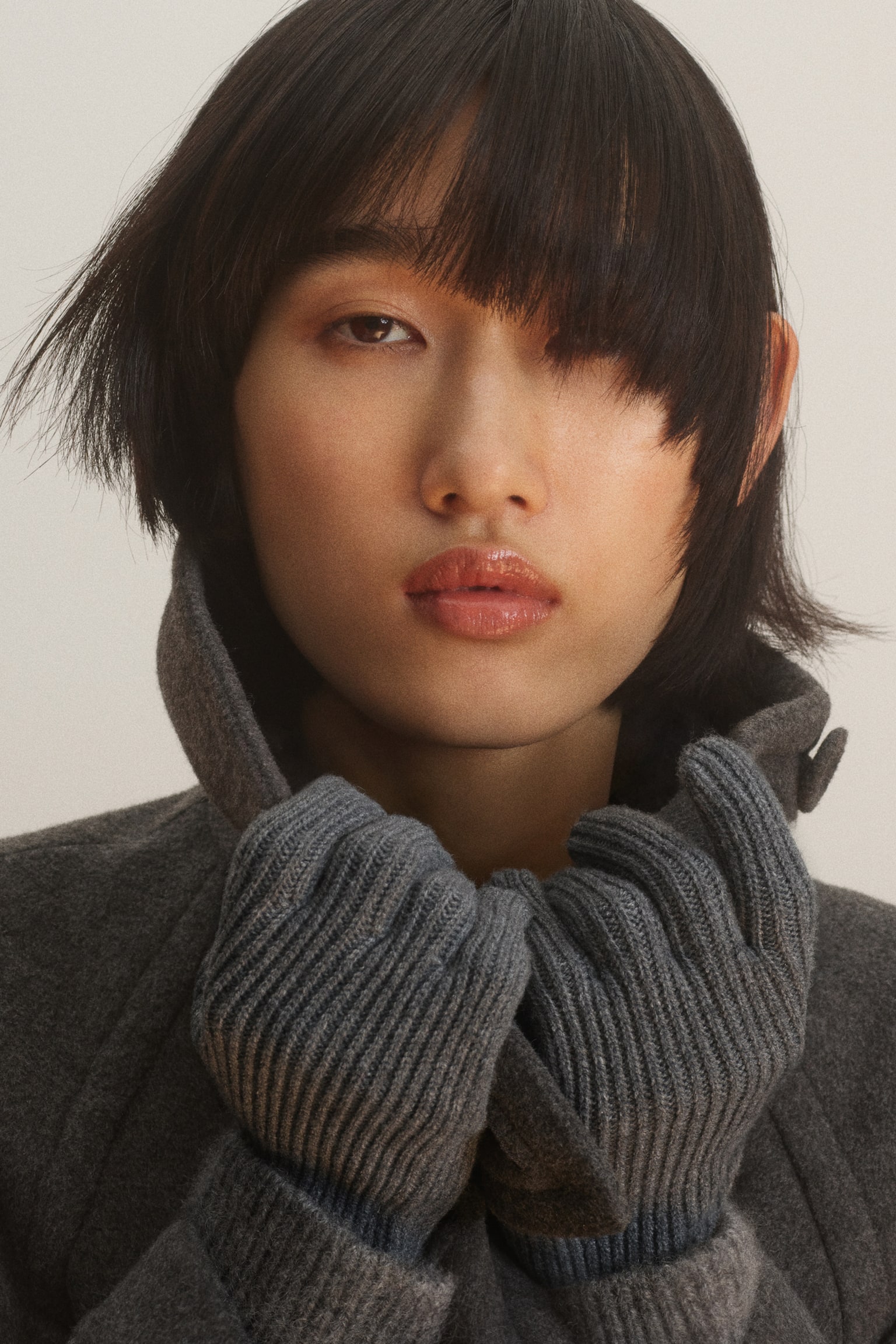 The model is wearing grey ribbed knit gloves and holding the high collar of a matching dark grey coat close to their face. - 1
