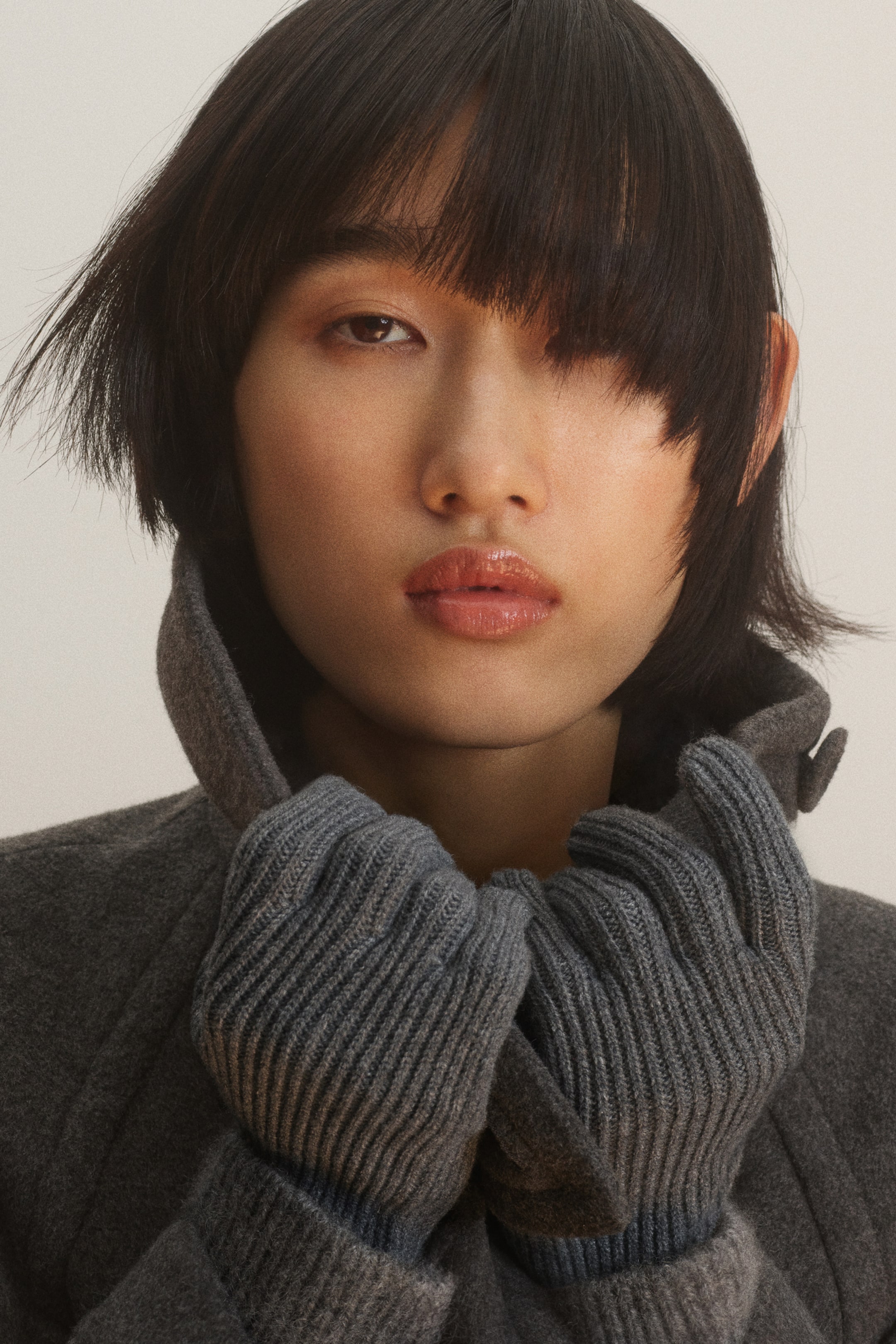Visualizza l'immagine ingrandita: La modella indossa guanti a maglia grigia a coste e tiene vicino al viso l’alta collo di un cappotto grigio scuro abbinato.