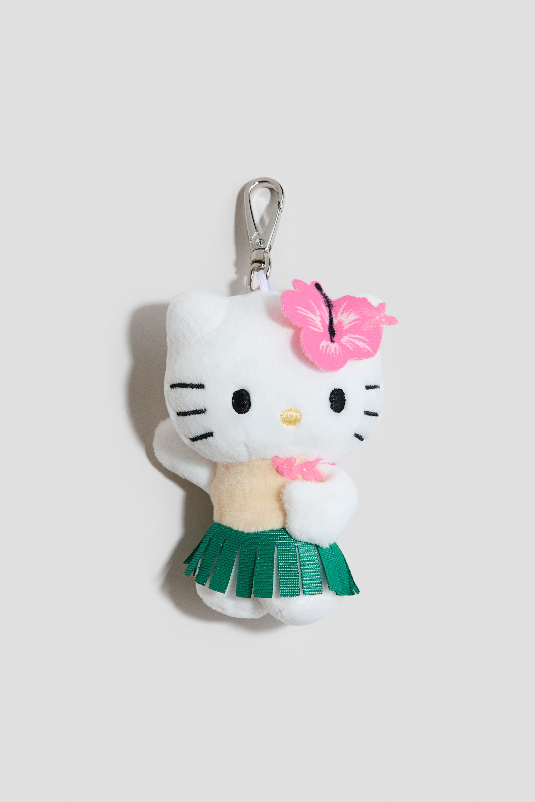 Ver imagen más grande: Un divertido colgante para bolso presenta un personaje de gato de peluche blanco, mostrado de frente, detallado con ojos negros, una nariz amarilla y bigotes negros. El personaje lleva una flor de hibisco rosa en la cabeza, una camiseta marrón claro, una falda de hula verde y un lei rosa, y se conecta a un mosquetón de metal plateado en la parte superior.