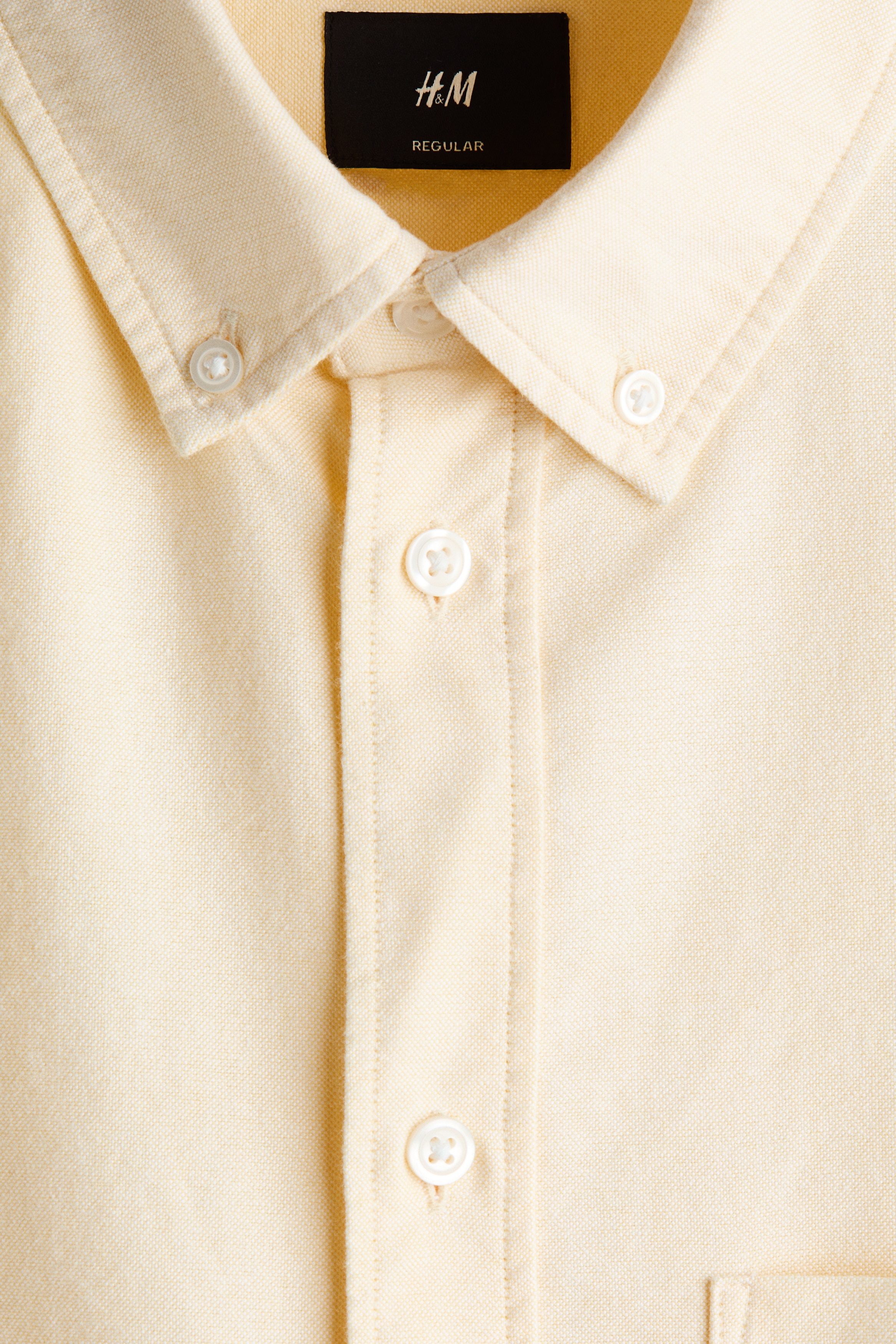 The model is wearing a pale yellow Oxford cotton shirt layered over a white T-shirt and tucked into dark trousers. The shirt features a button-down collar, a classic button-front closure, a single chest pocket, and long sleeves rolled up at the cuffs.